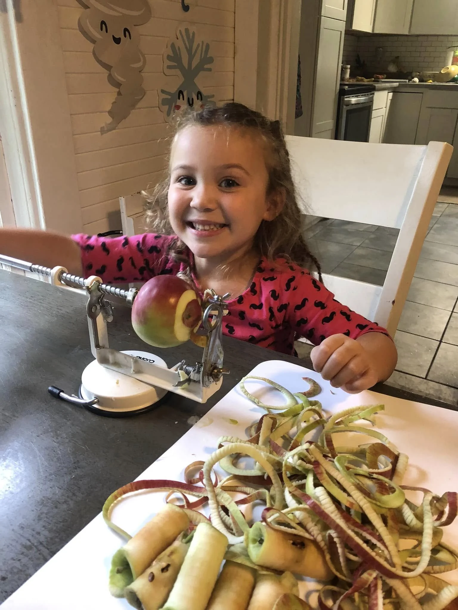 A young girl with curly hair, smiling, sitting at a table with a cutting board full of peeled and sliced apples and spiralized apple peels. There is a small apple peeler tool on the table and a decorative wall with a happy face and a tree in the back