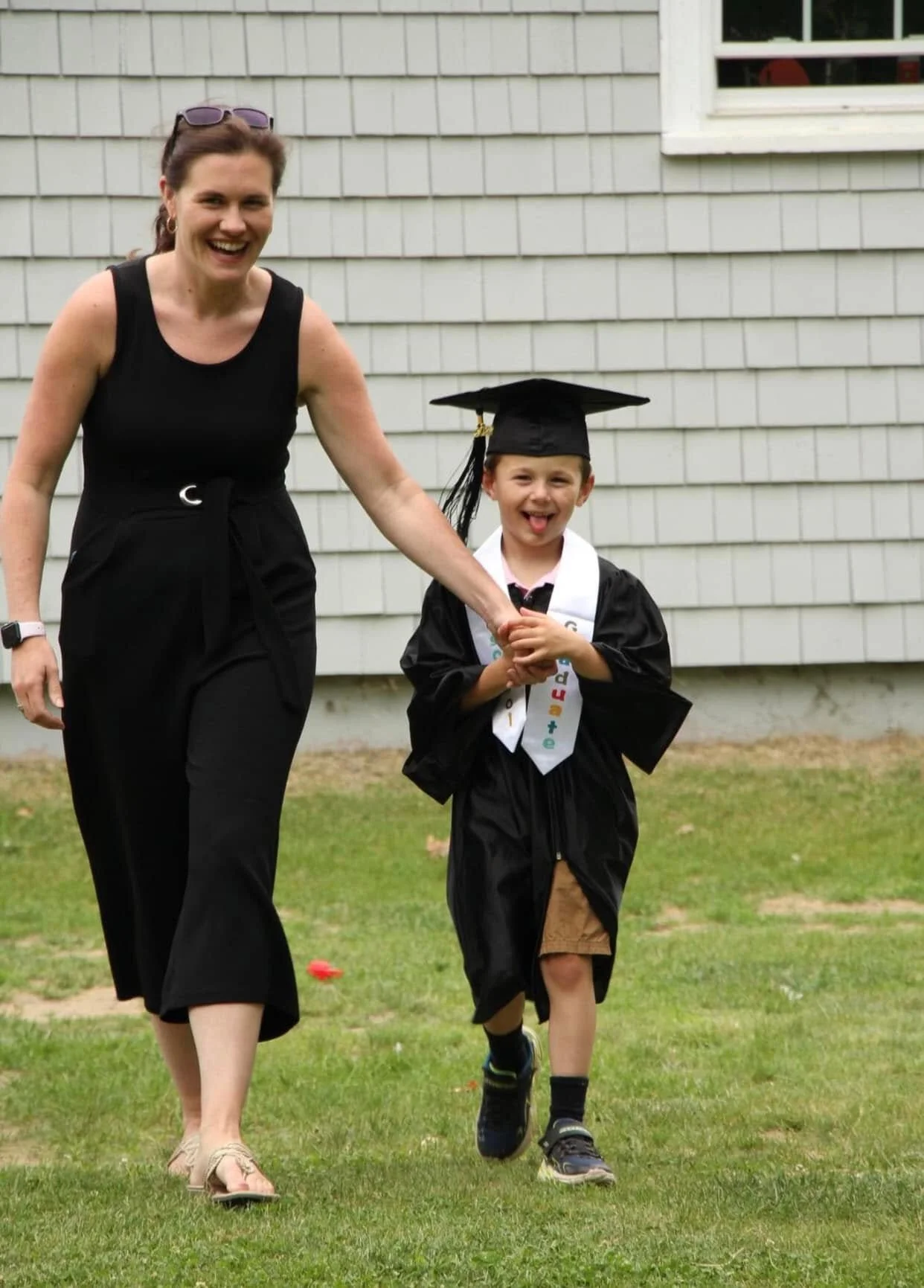 A woman and a young boy are outdoors, walking on grass in front of a house with gray siding. The woman is wearing a black sleeveless top and black pants, smiling, and holding the boy's hand. The boy is wearing a graduation cap and gown with a colorfu