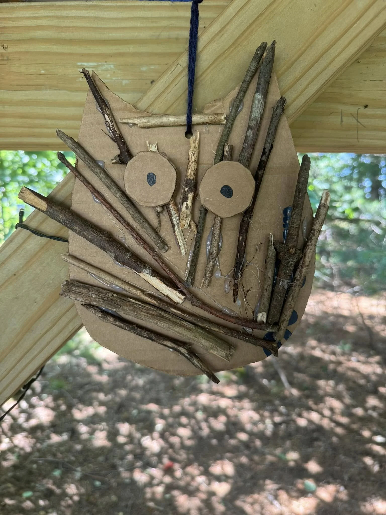 A handmade wooden wall hanging featuring two circular cutouts with black dots in the center, various sticks and twigs attached, and a blue string hanging from the top, mounted on a wooden surface outdoors.