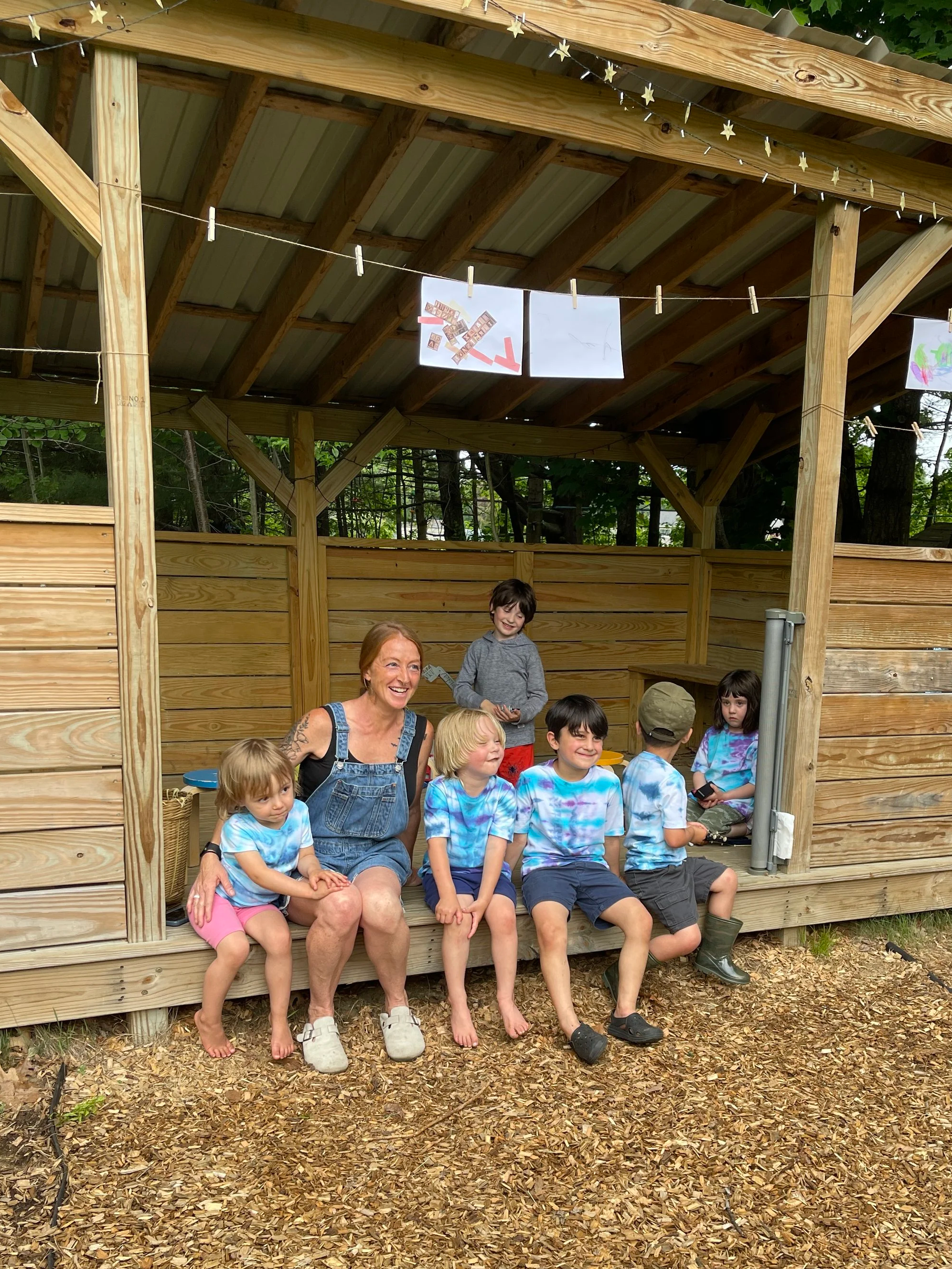 A group of children and one adult sitting and standing in front of a wooden outdoor shelter with a metal roof. The children are wearing tie-dye shirts, and the adult is smiling. The shelter is decorated with string lights and hanging signs. The ground is covered with wood chips, and trees are visible in the background.
