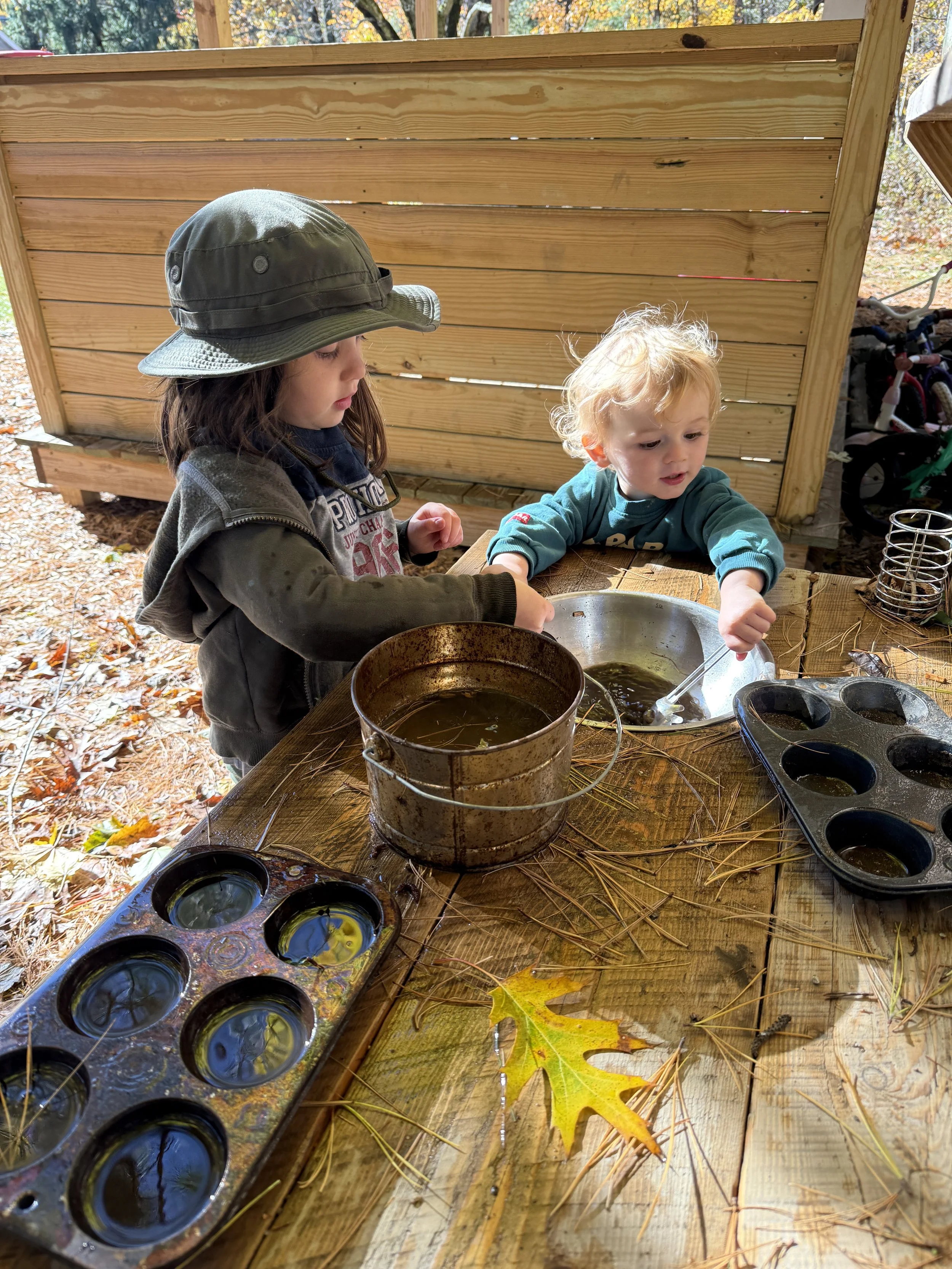 Two children, one girl with a hat and jacket, and a younger boy with curly blonde hair, playing with water in a large metal container and a muffin tin on a wooden outdoor table surrounded by fallen autumn leaves.