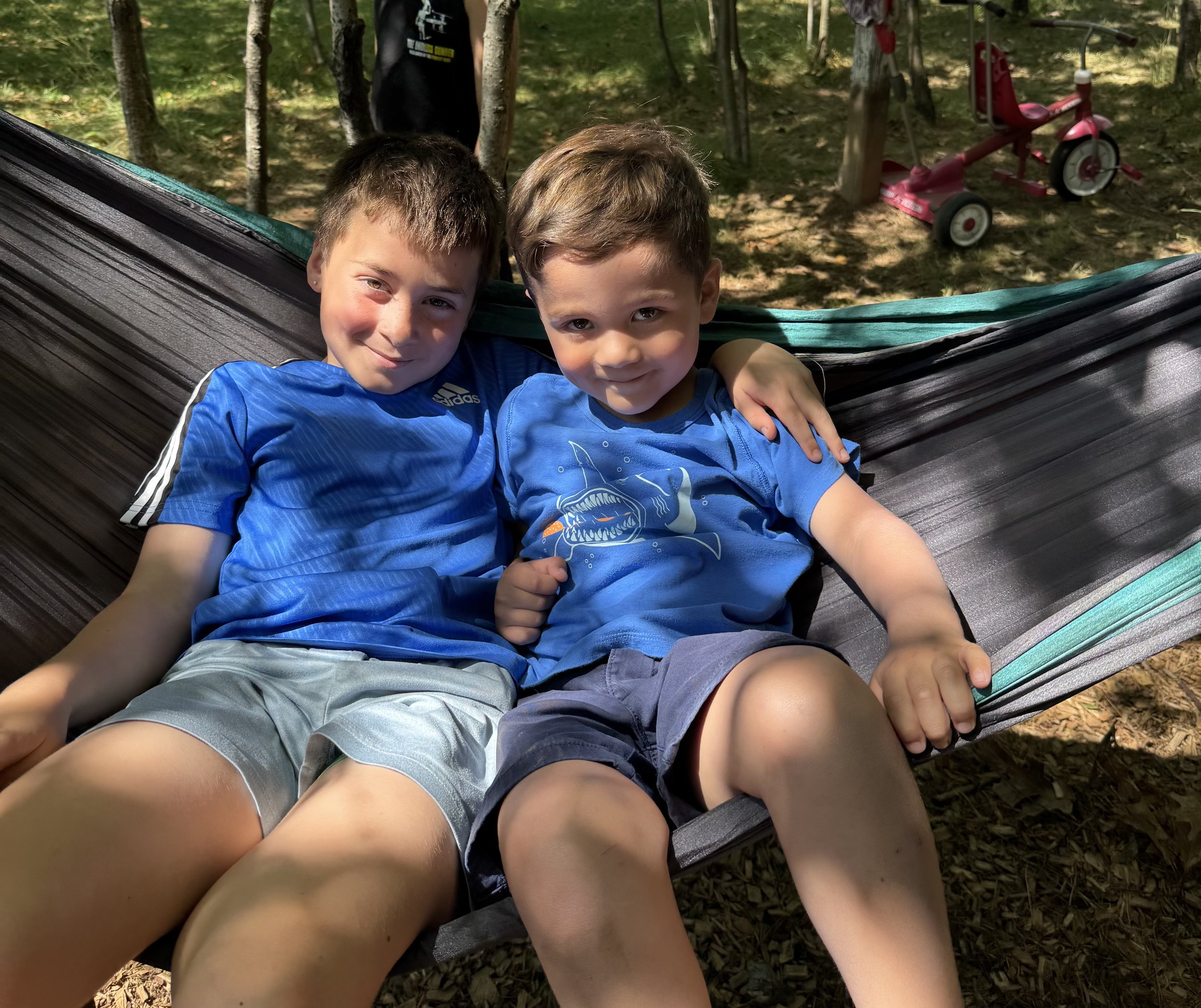 Two young boys sitting on a hammock in a wooded outdoor area, with a pink tricycle in the background.