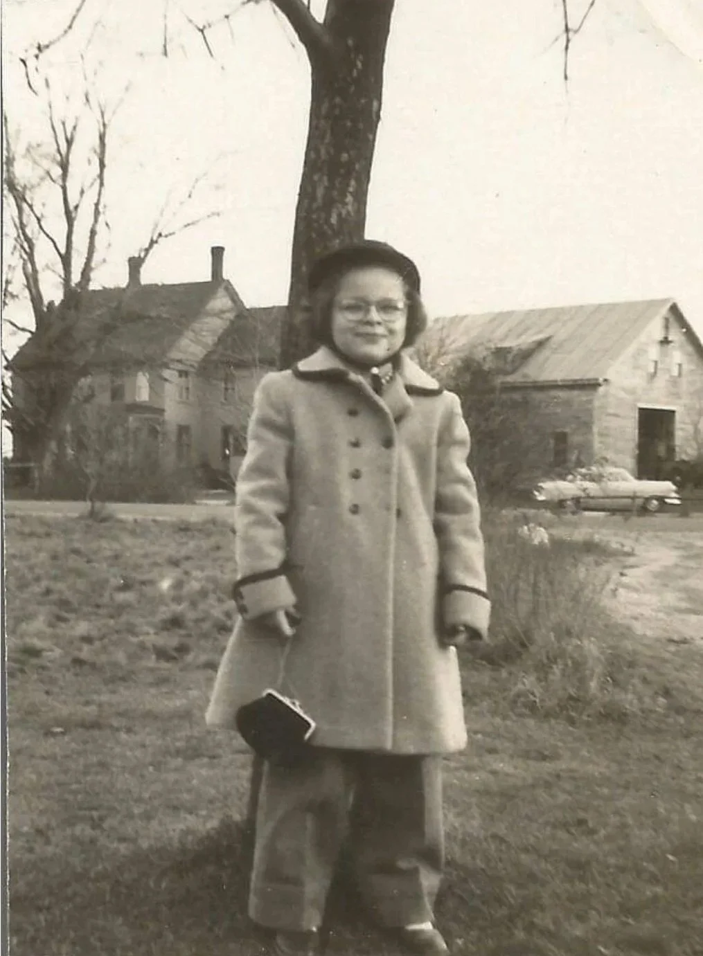 A young girl wearing glasses and a long coat standing outdoors in front of a large tree, with houses and a car in the background, holding a small purse.
