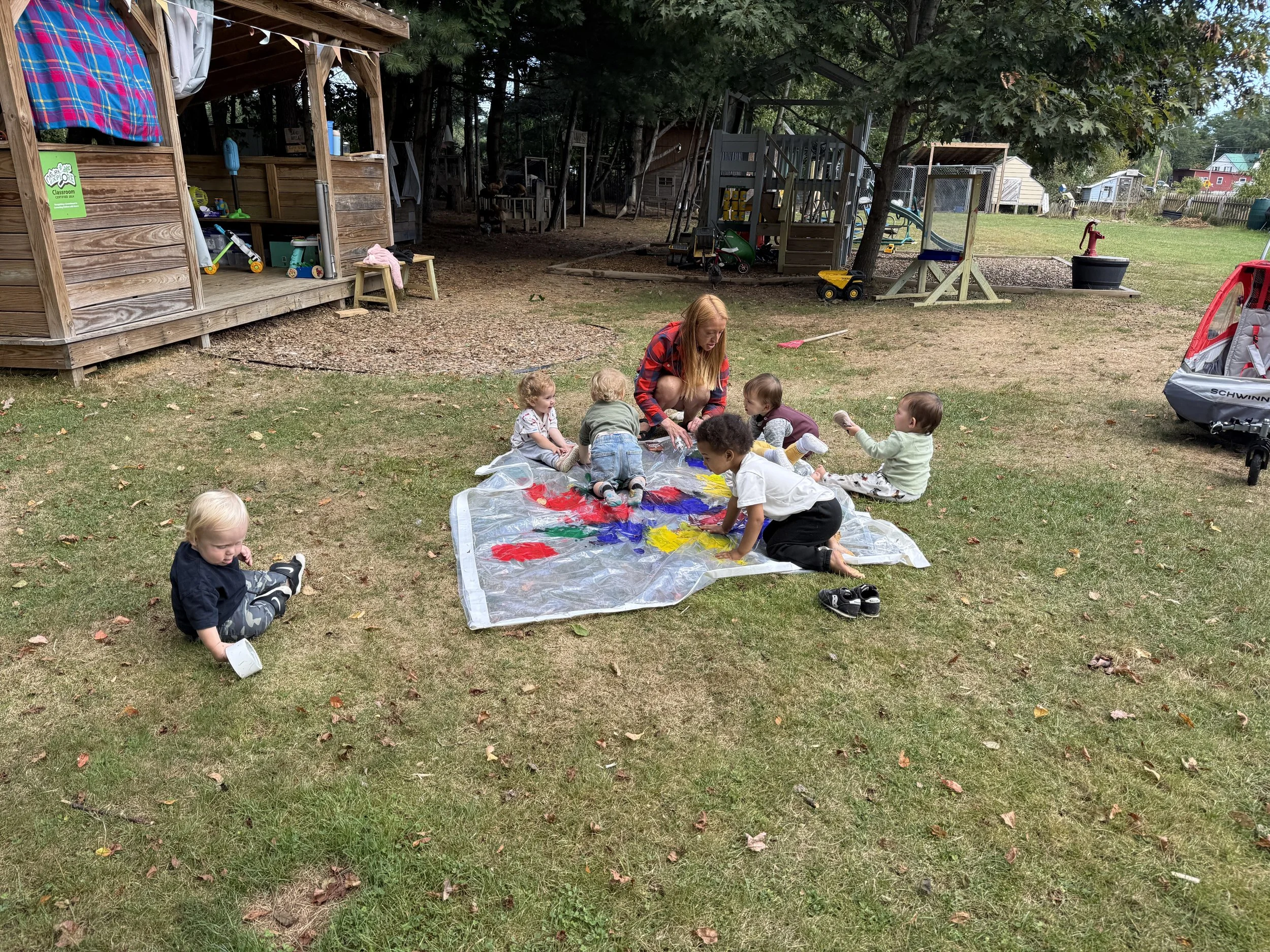 Children sitting and playing on a large transparent sheet with colorful paint splotches, under a tree in a backyard with play structures, toys, and grass.