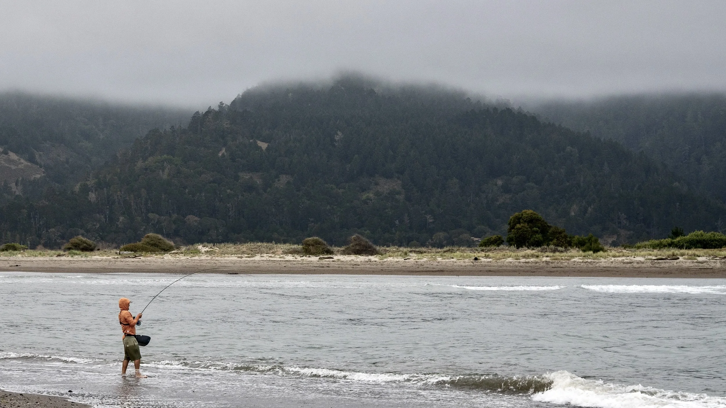 A person wearing a hoodie and shorts stands in the water while fishing, with a mountain range covered in fog in the background.
