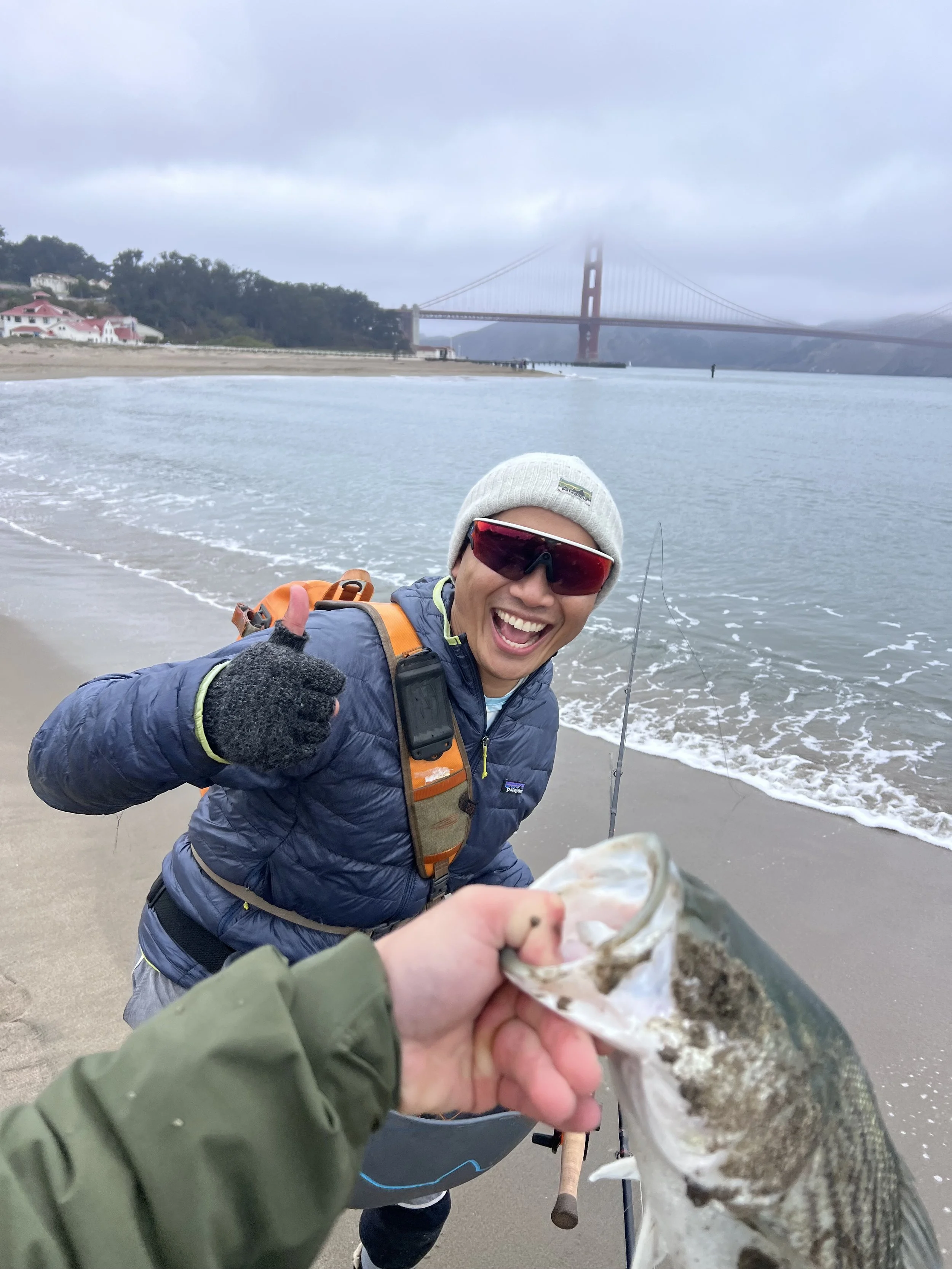 Pure excitement and  a thumbs-up while holding a fish on a beach, with the Golden Gate Bridge partially shrouded in fog in the background.