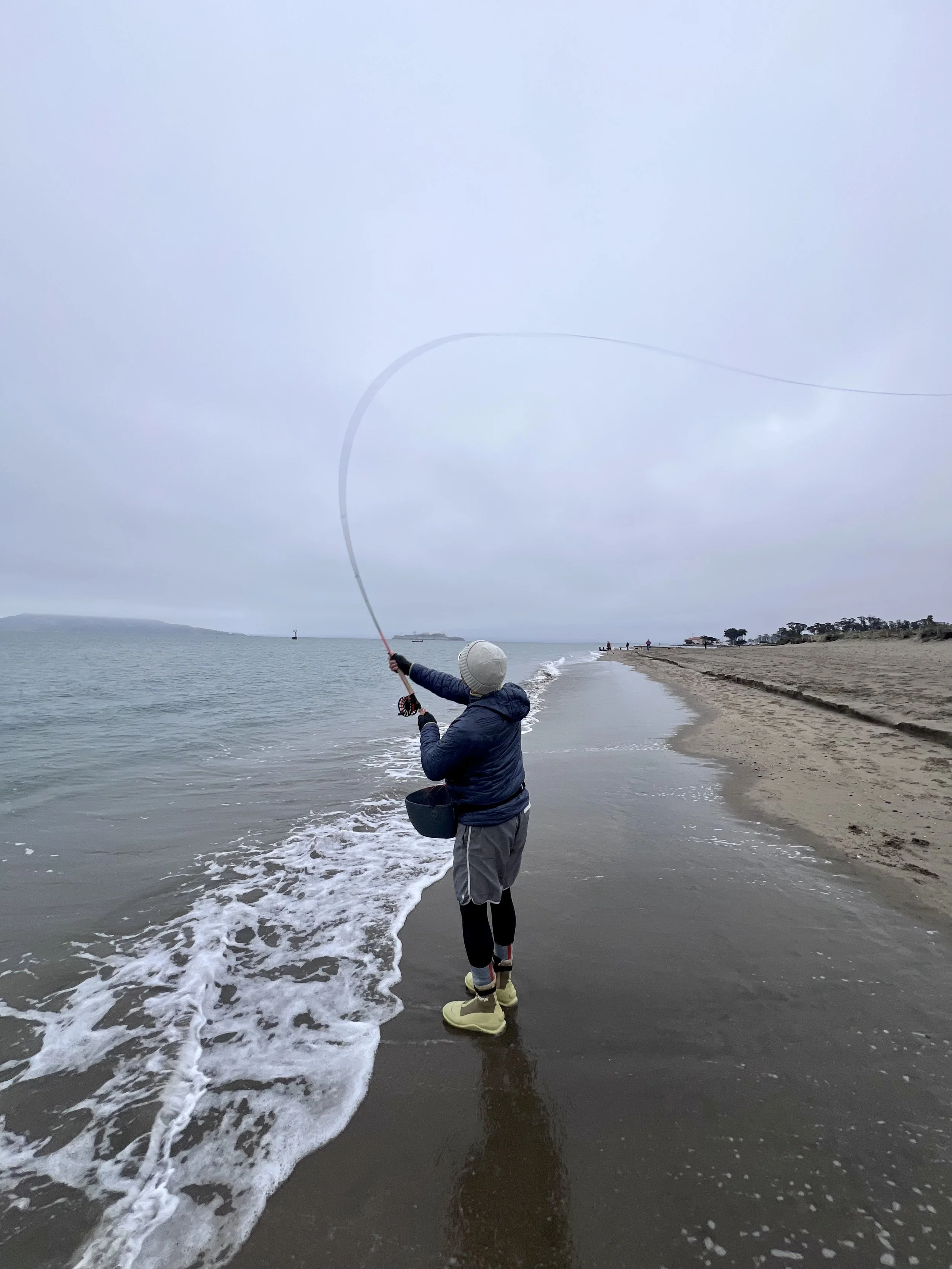 A person standing on a beach near the water's edge, casting a fishing line into the ocean on a cloudy day.