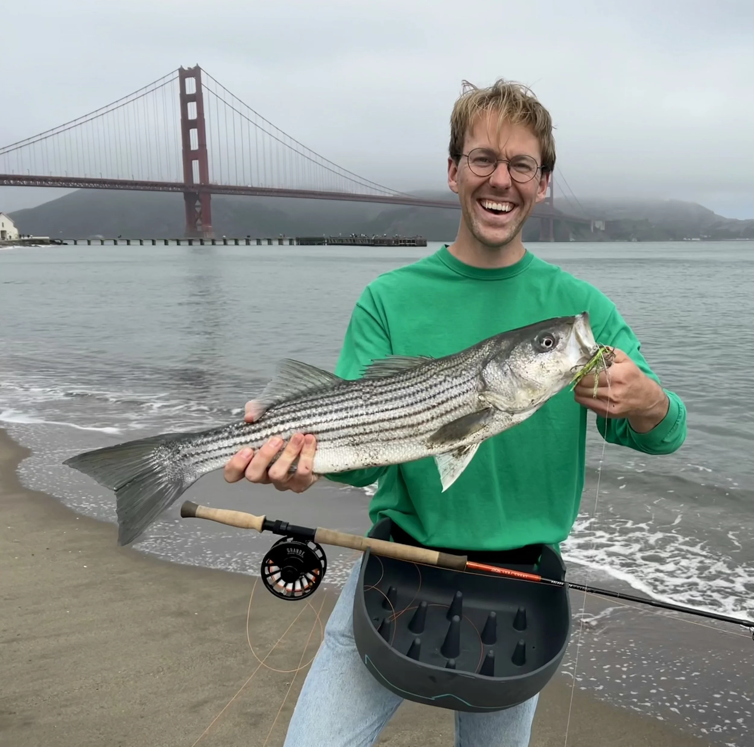 A man with glasses, smiling, wearing a green shirt, holding a fish on a beach with the Golden Gate Bridge in the background under overcast skies.