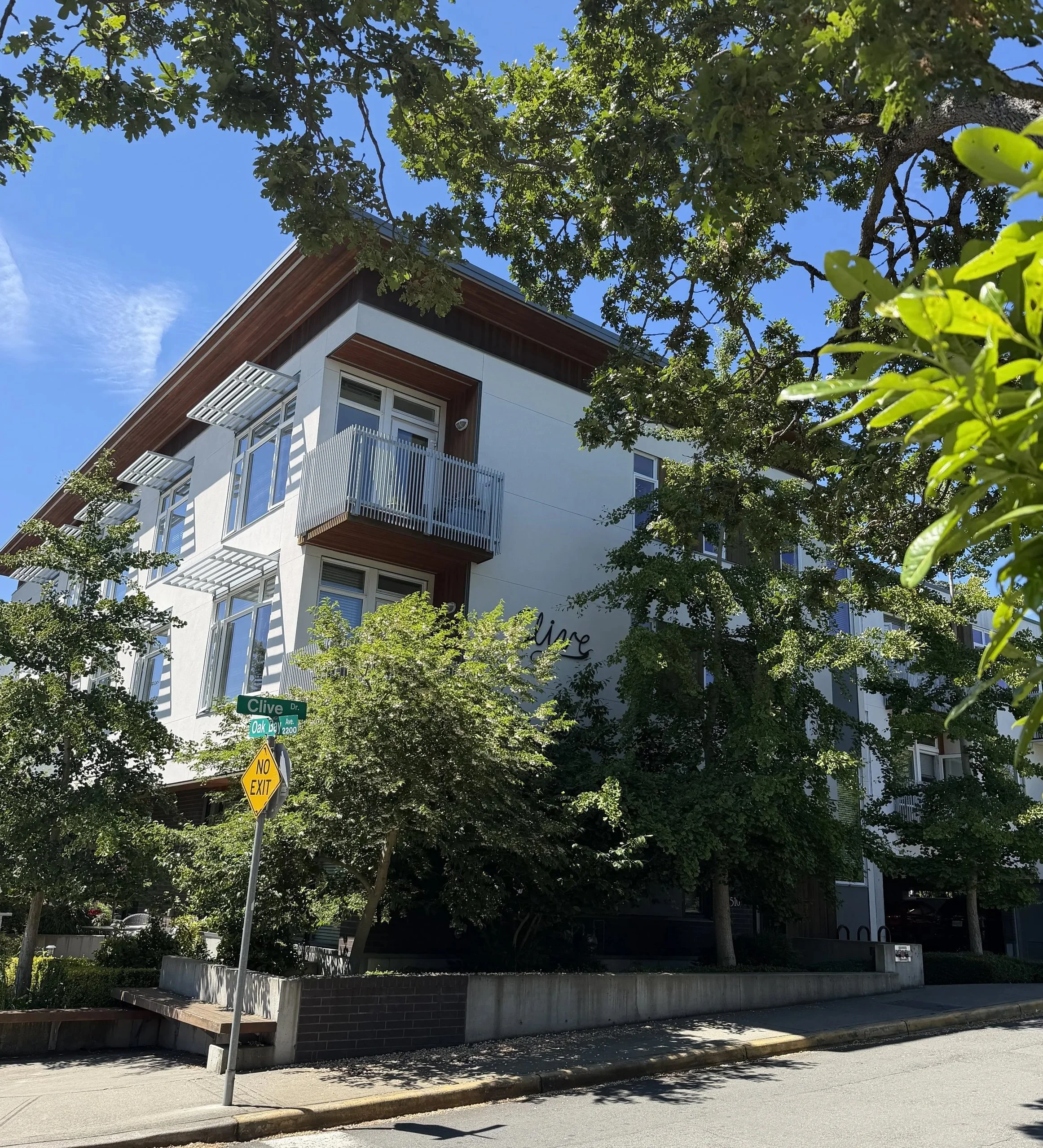 A modern multi-story apartment building with balconies, surrounded by green trees, located at the intersection of Clive Drive and Oak Bay Avenue. The sky is clear and blue.