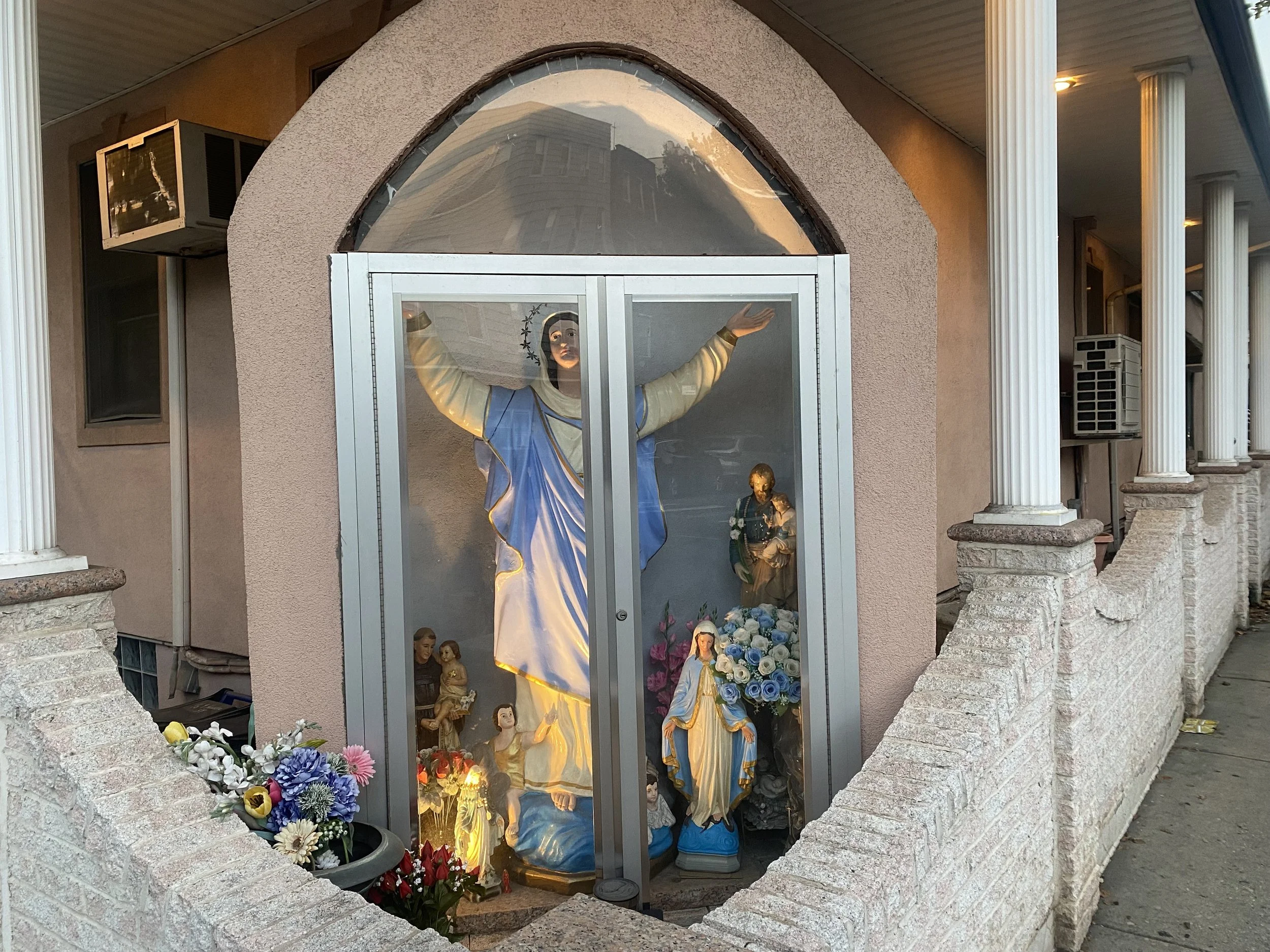Religious shrine featuring a statue of the Virgin Mary with arms raised. The shrine is enclosed in a glass case with flowers and smaller religious figures at the base. The structure has a stucco exterior with arched glass window and is supported by white columns.