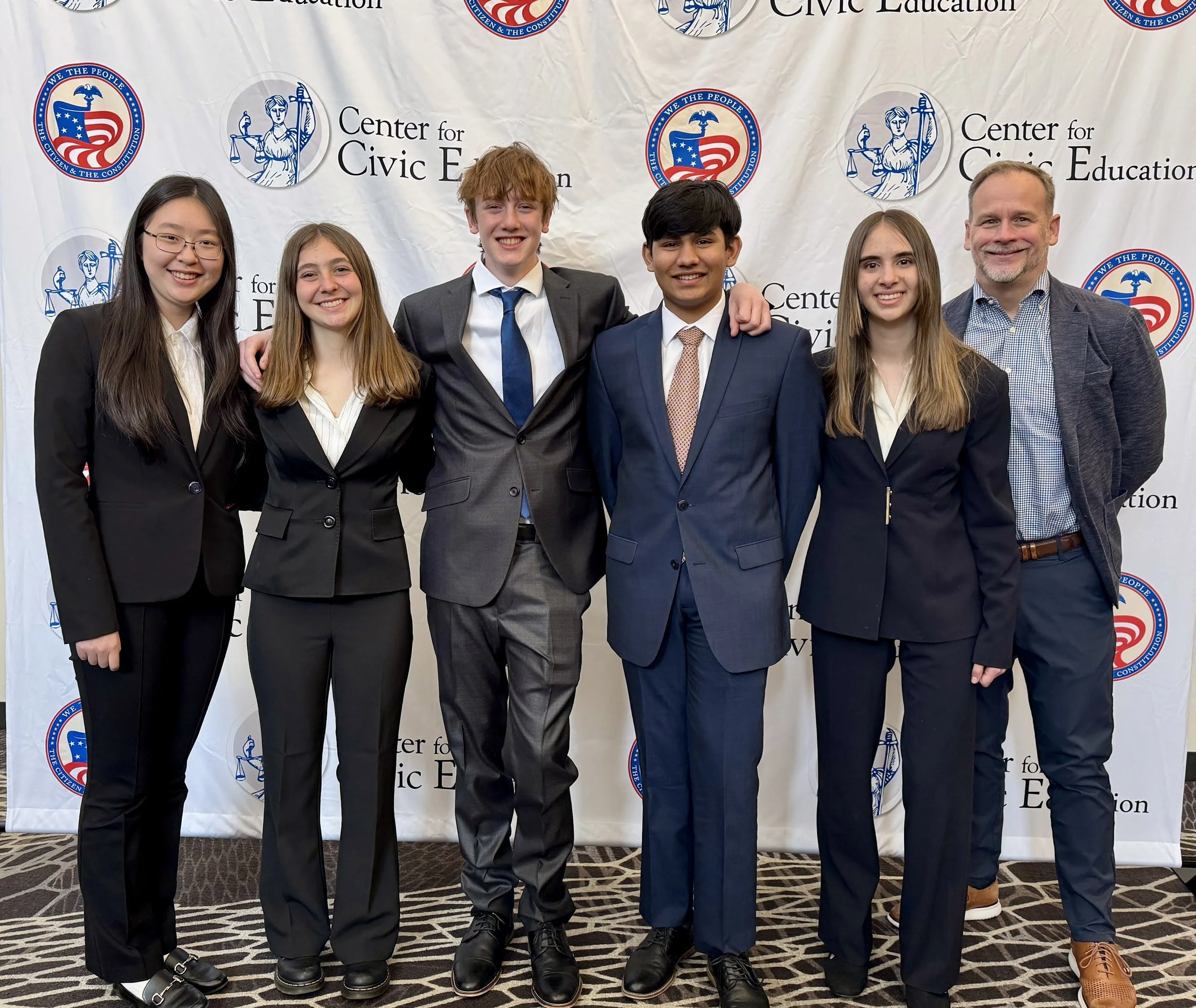 A group of six young people, five women and one man, standing in front of a backdrop with logos for the Center for Civic Education and the We the People emblem. They are dressed in formal attire, smiling, with arms around each other.