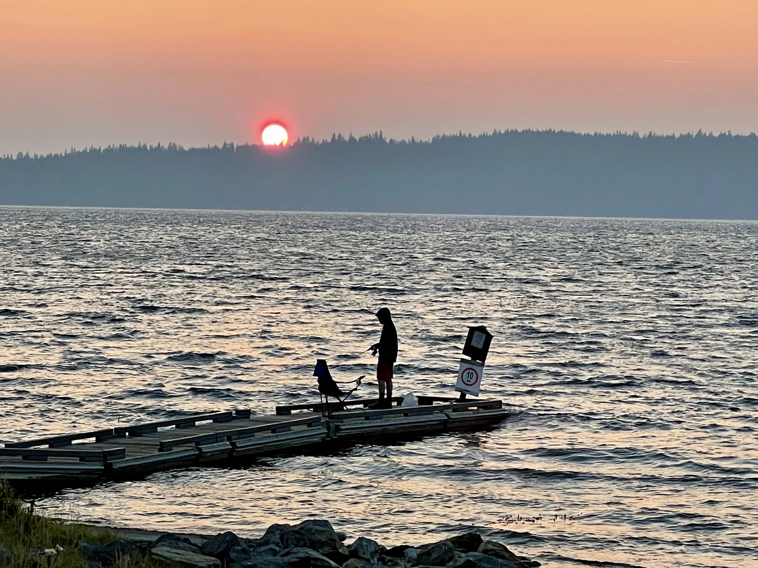 Sunset over a body of water with a silhouetted person fishing on a dock and a seagull next to them.