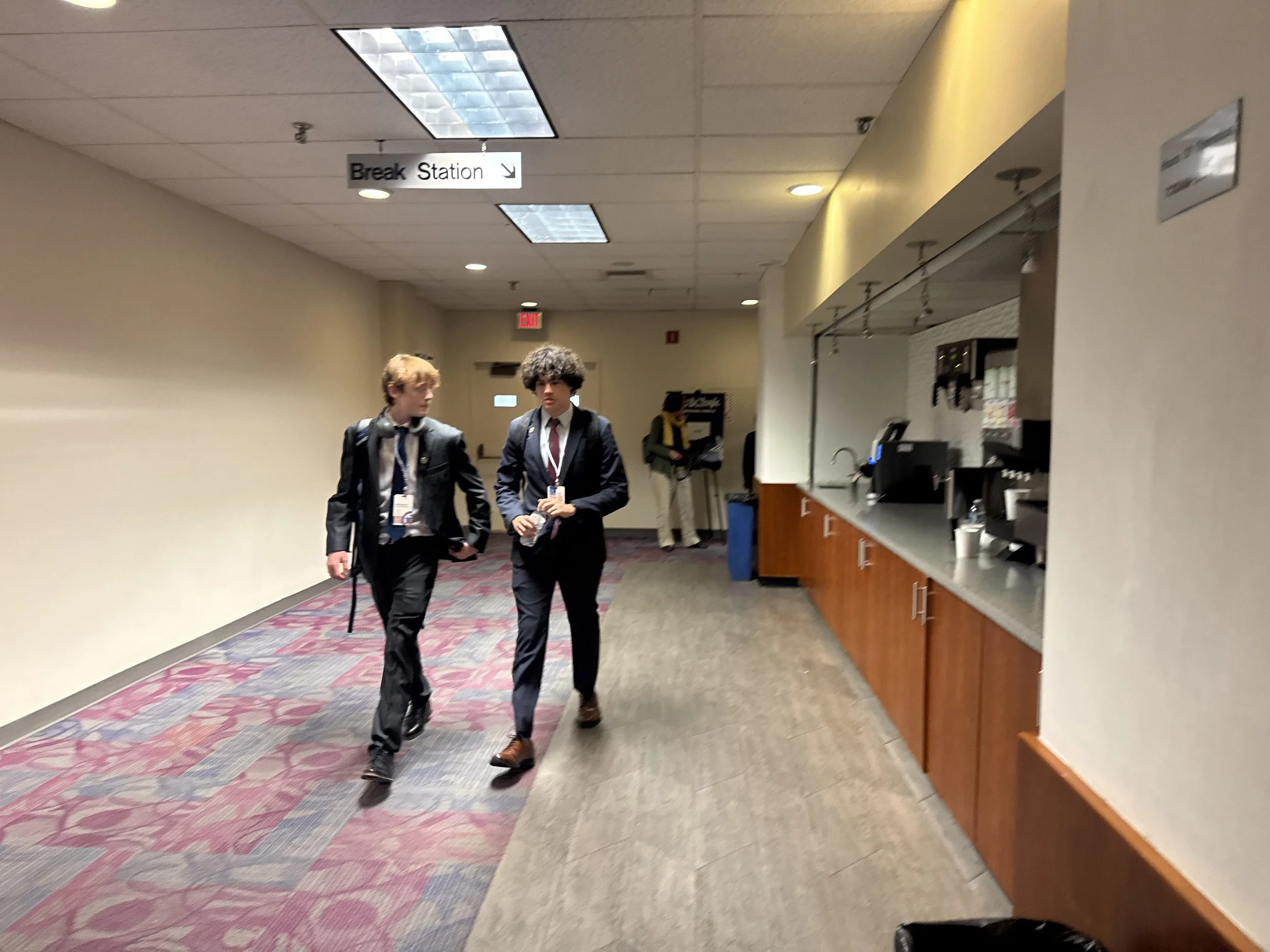 Two young men walking down a corridor in a building, likely a conference or school setting. They are wearing suits and carry backpacks, with one also holding a water bottle. There are ceiling lights, a 'Break Station' sign hanging from the ceiling, and a cafeteria or service area on the right side.
