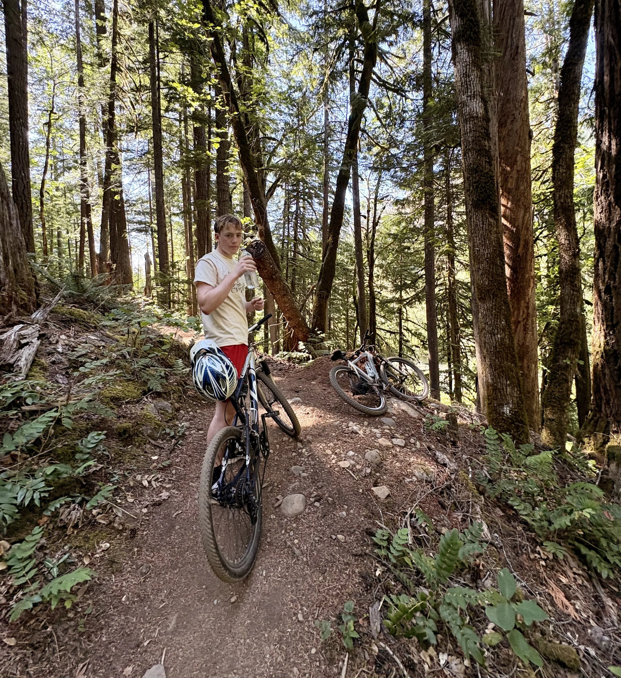 A young person in a forested area, taking a break during a bike ride, holding a water bottle and surrounded by bicycles on a dirt trail.