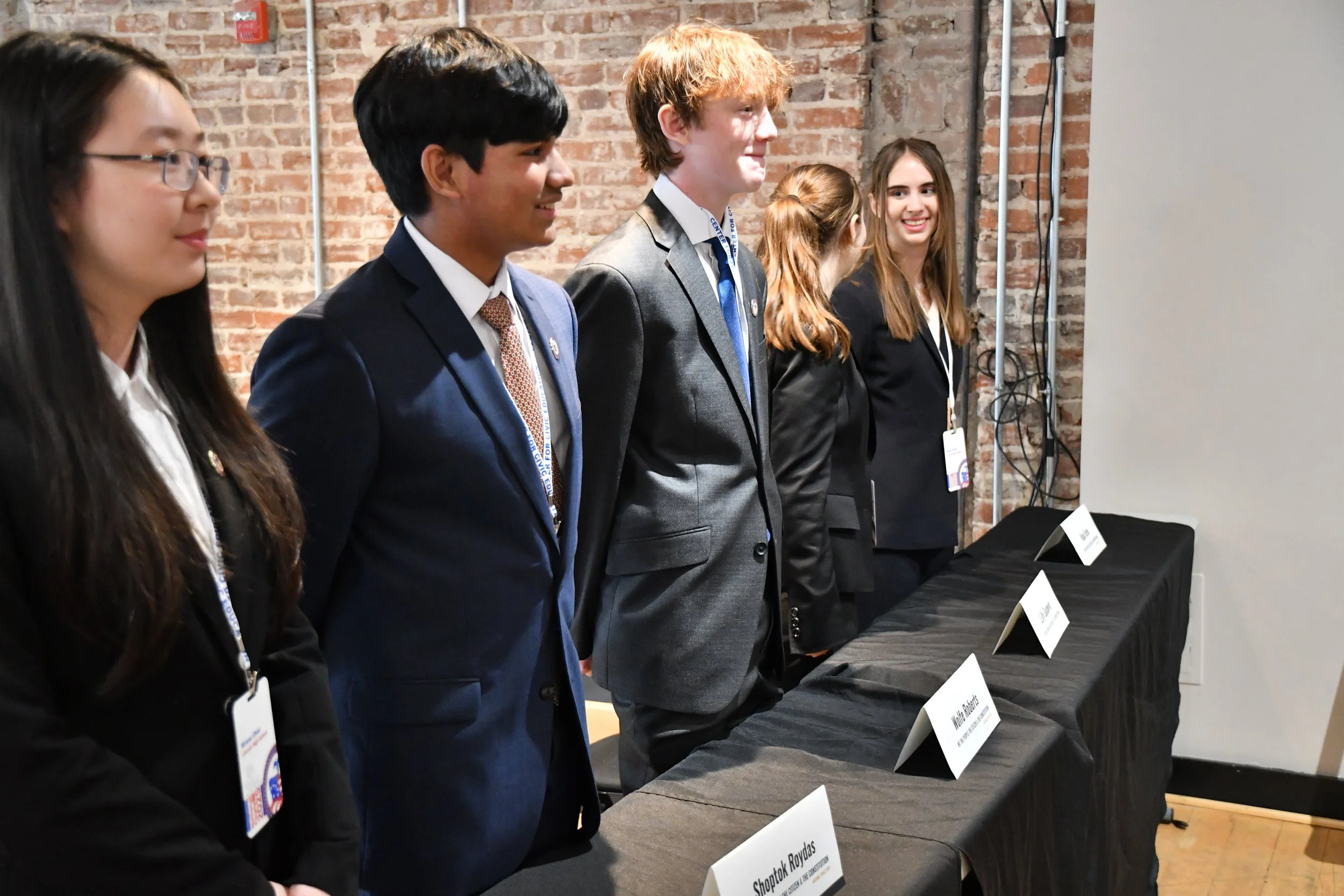 A group of young people dressed in business attire standing in a line at a conference, event, or competition with name cards in front of them on a table.