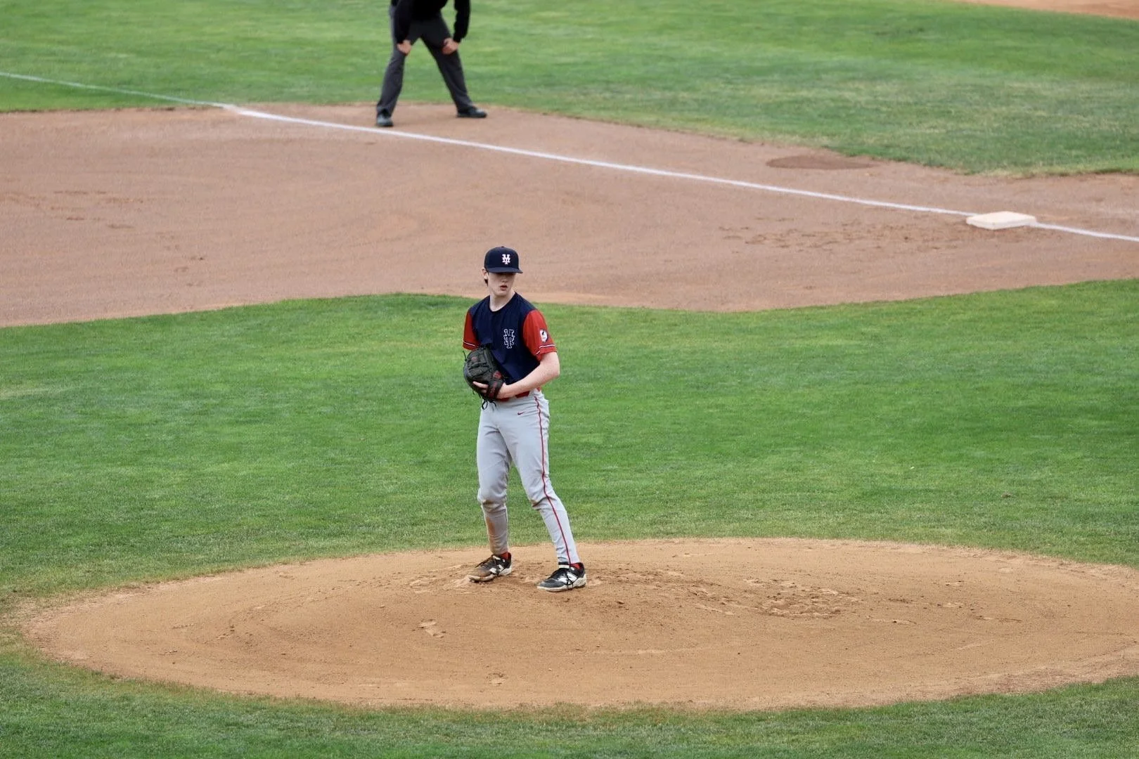 A young baseball player stands on the pitcher's mound holding a baseball glove, with a baseball field and an umpire in the background.
