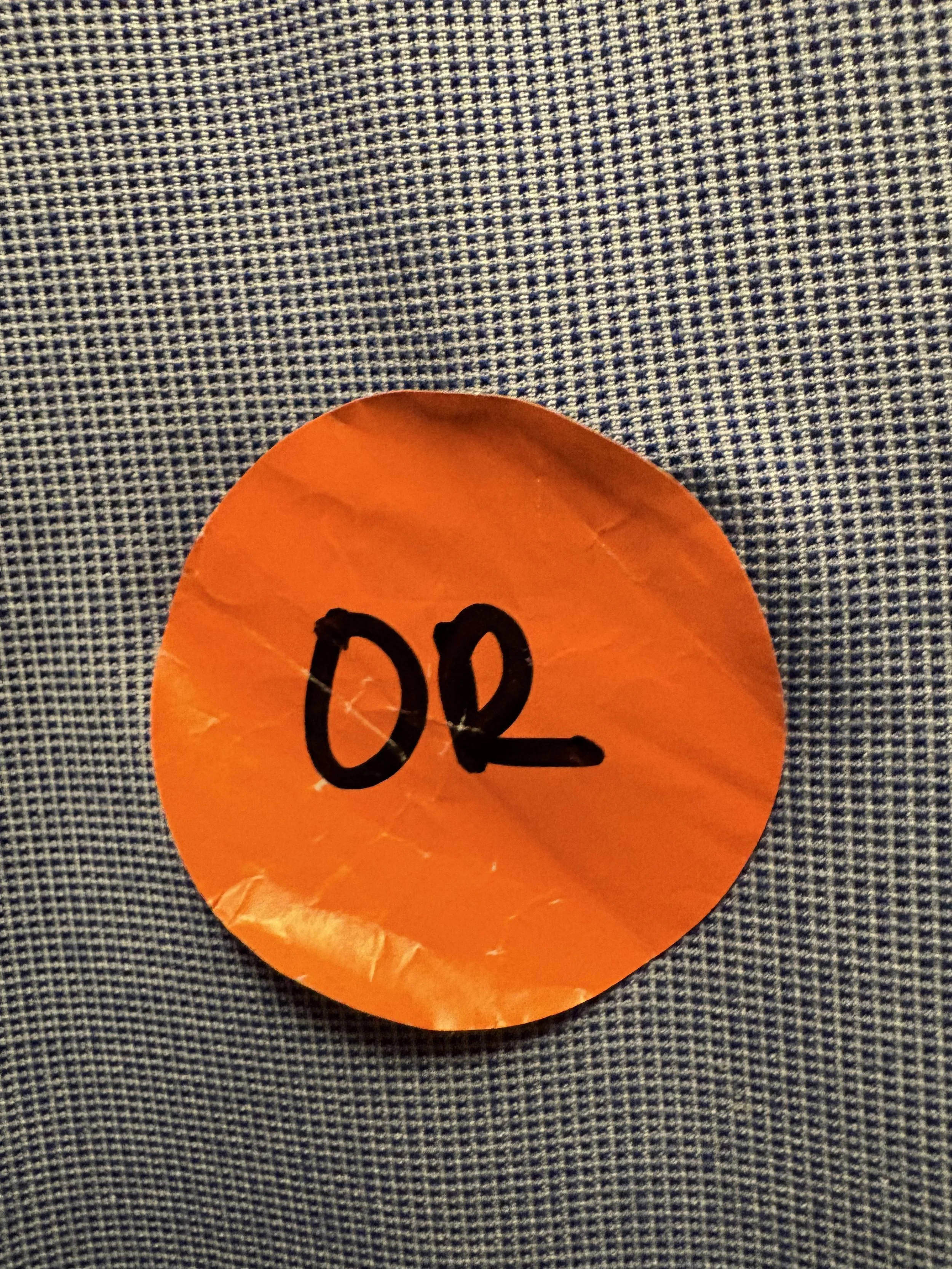 Orange paper sticker with black handwritten letters spelling 'OOR' placed on a dark gray checkered fabric surface.