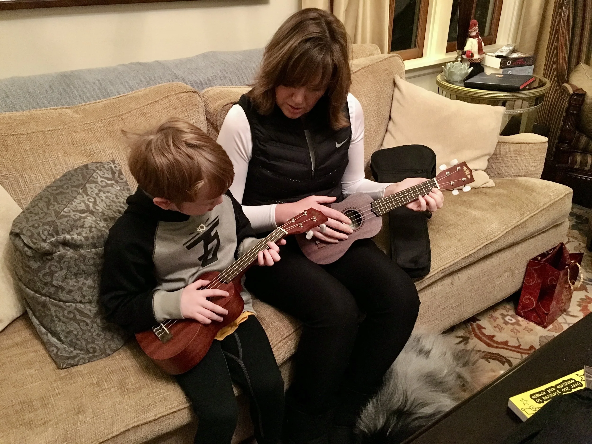A woman and a young boy sitting on a couch, both playing small ukuleles. The scene is in a cozy living room with a window, some cushions, a side table with a snowman and other objects, and patterned carpet.