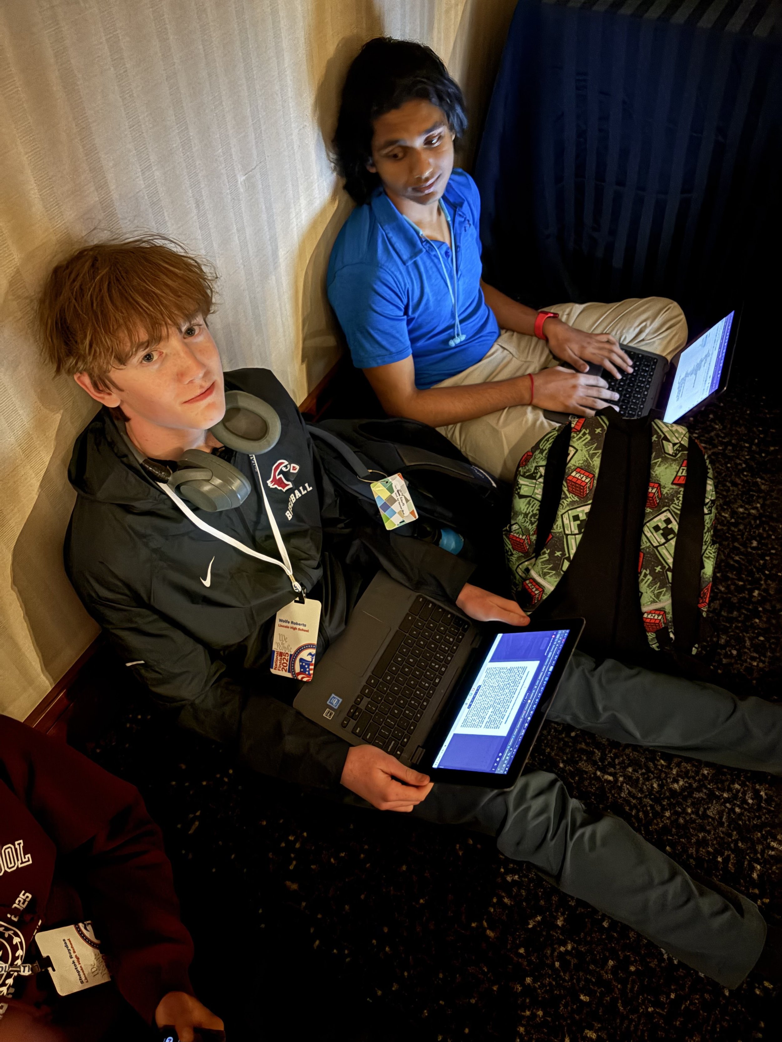Two young men seated against a wall with laptops, one with red hair and headphones around neck, the other with dark hair and a red wristband, both working on their computers.