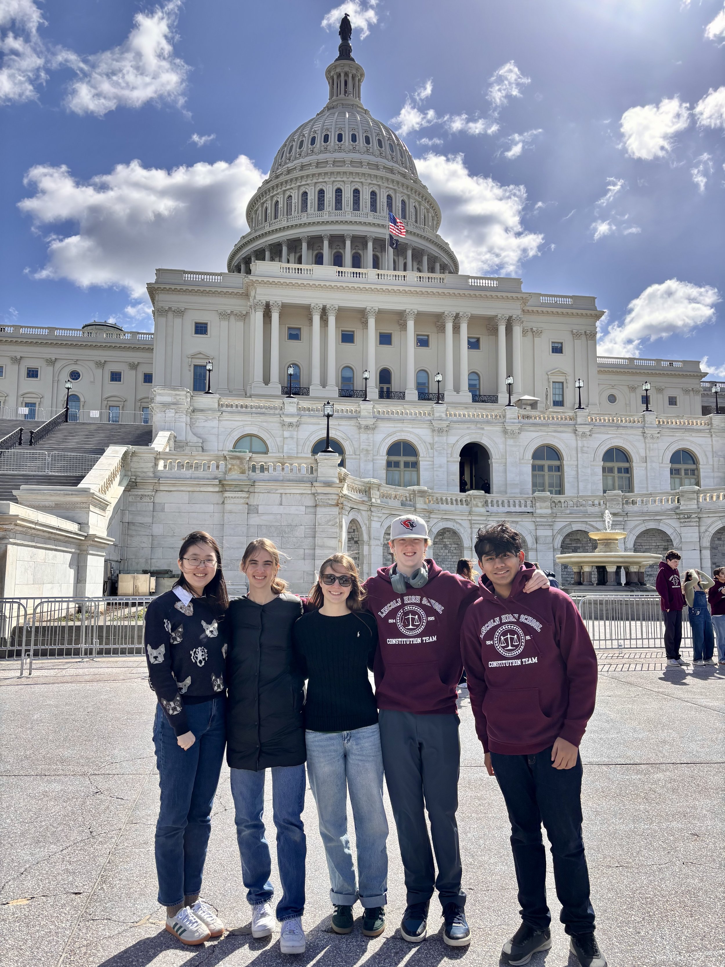 Five young people standing together in front of the United States Capitol building in Washington D.C. on a sunny day. They are smiling and appear to be happy, with three males and two females in the group.