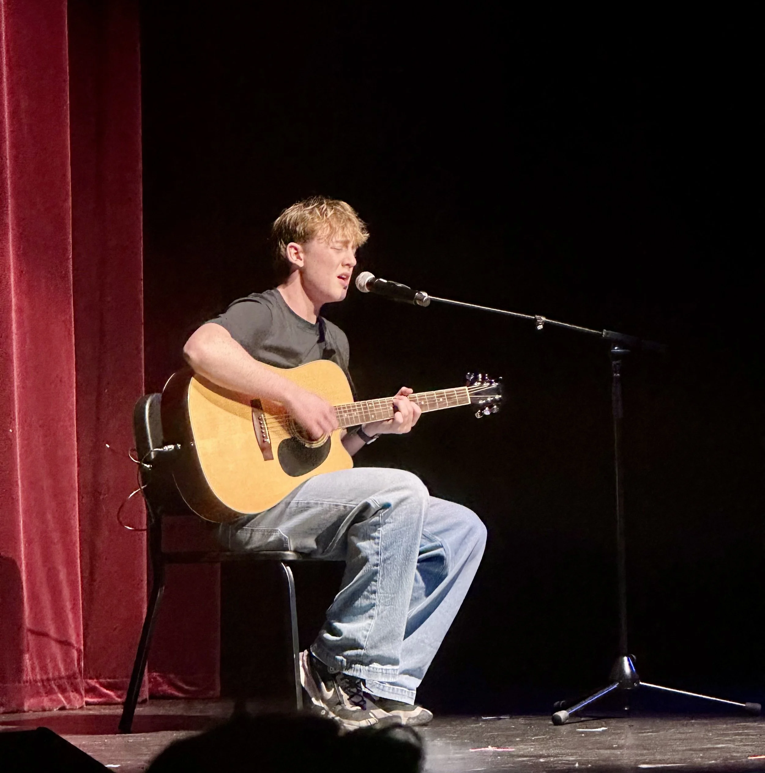 A young man singing into a microphone and playing an acoustic guitar on a stage with red curtains and a black backdrop.