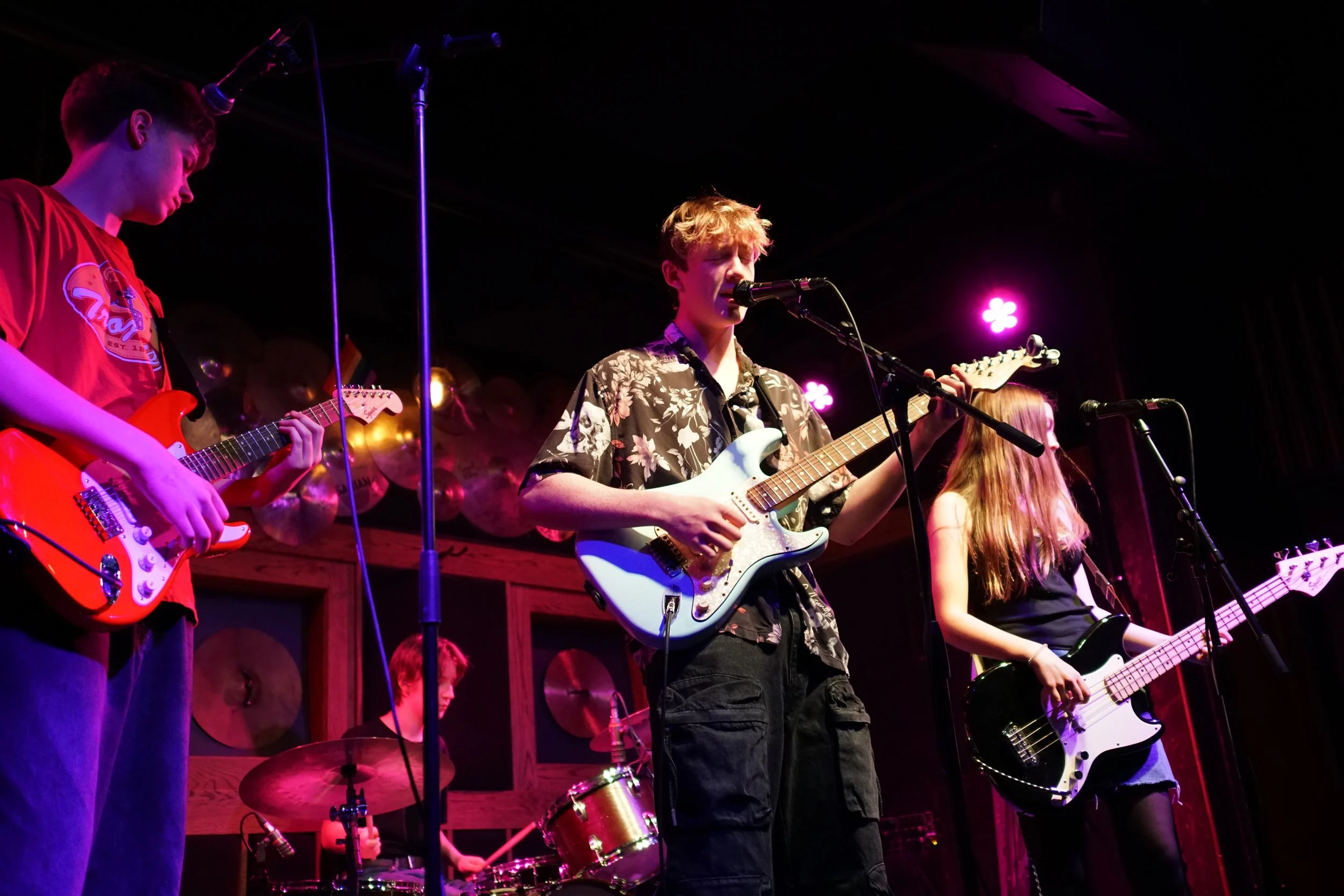 Four musicians, three females and one male, performing on a stage at night. They are playing guitars and drums, with purple and pink stage lighting, a dark background, and a wooden wall behind them.