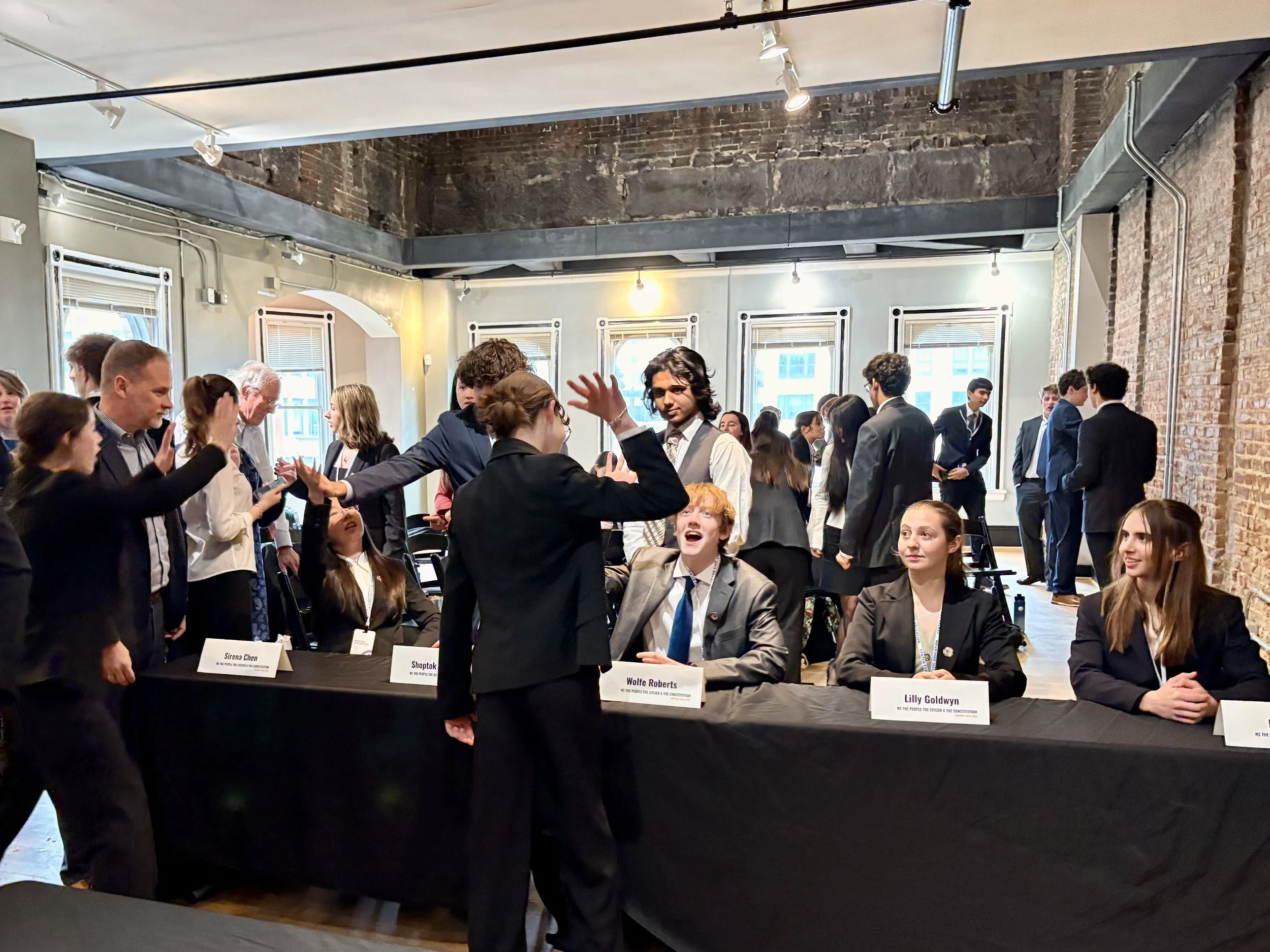Group of people at an indoor event, with some people sitting at a table with nameplates, and others standing and interacting. The setting has exposed brick walls and large windows.