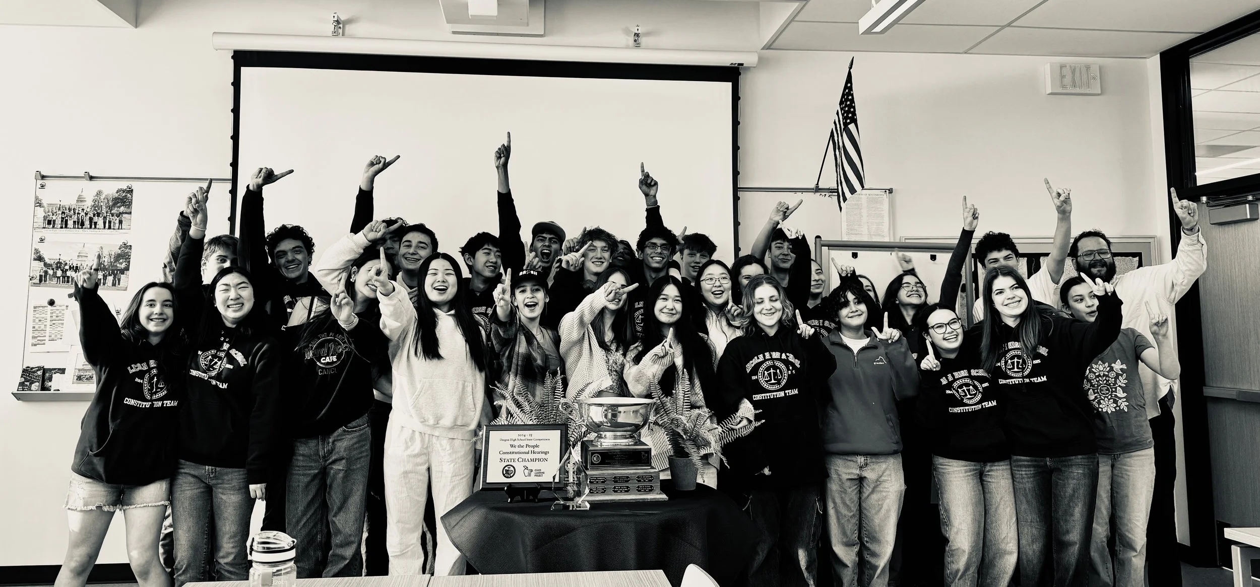A group of students and a teacher celebrating a victory or achievement in a classroom, some students wearing hoodies with a logo, standing behind a table with a trophy and a plaque, all smiling and raising their hands.