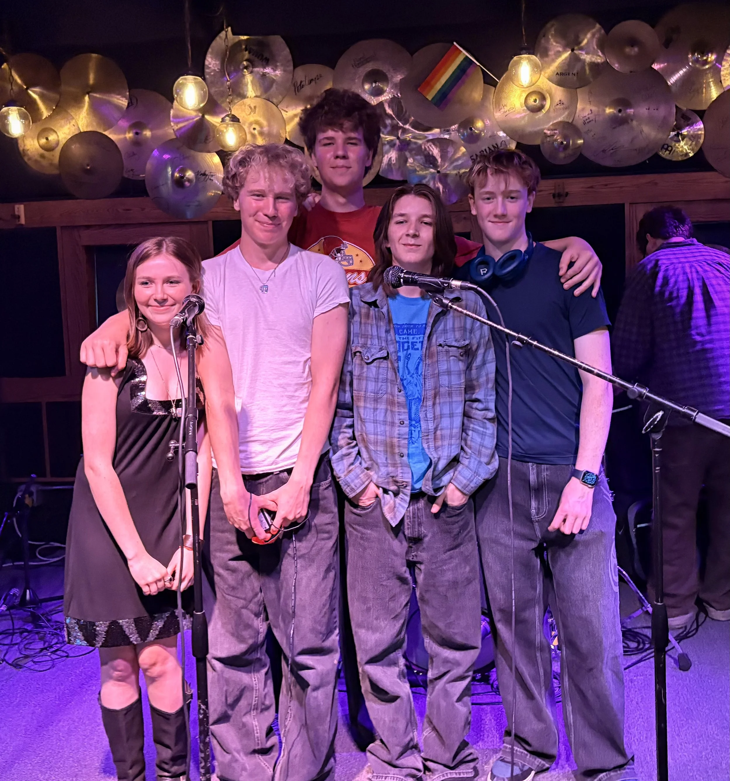 A group of five young people standing together on a stage with a background of cymbals and purple lighting. They are smiling and some have their arms around each other. There are microphones in front of them, and one audience member is visible in the background. The group appears to be in a music or performance setting.