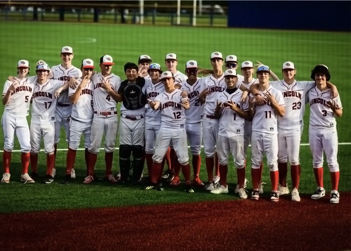 Group of young male baseball players in uniform posing on a baseball field with green grass and a fence in the background.