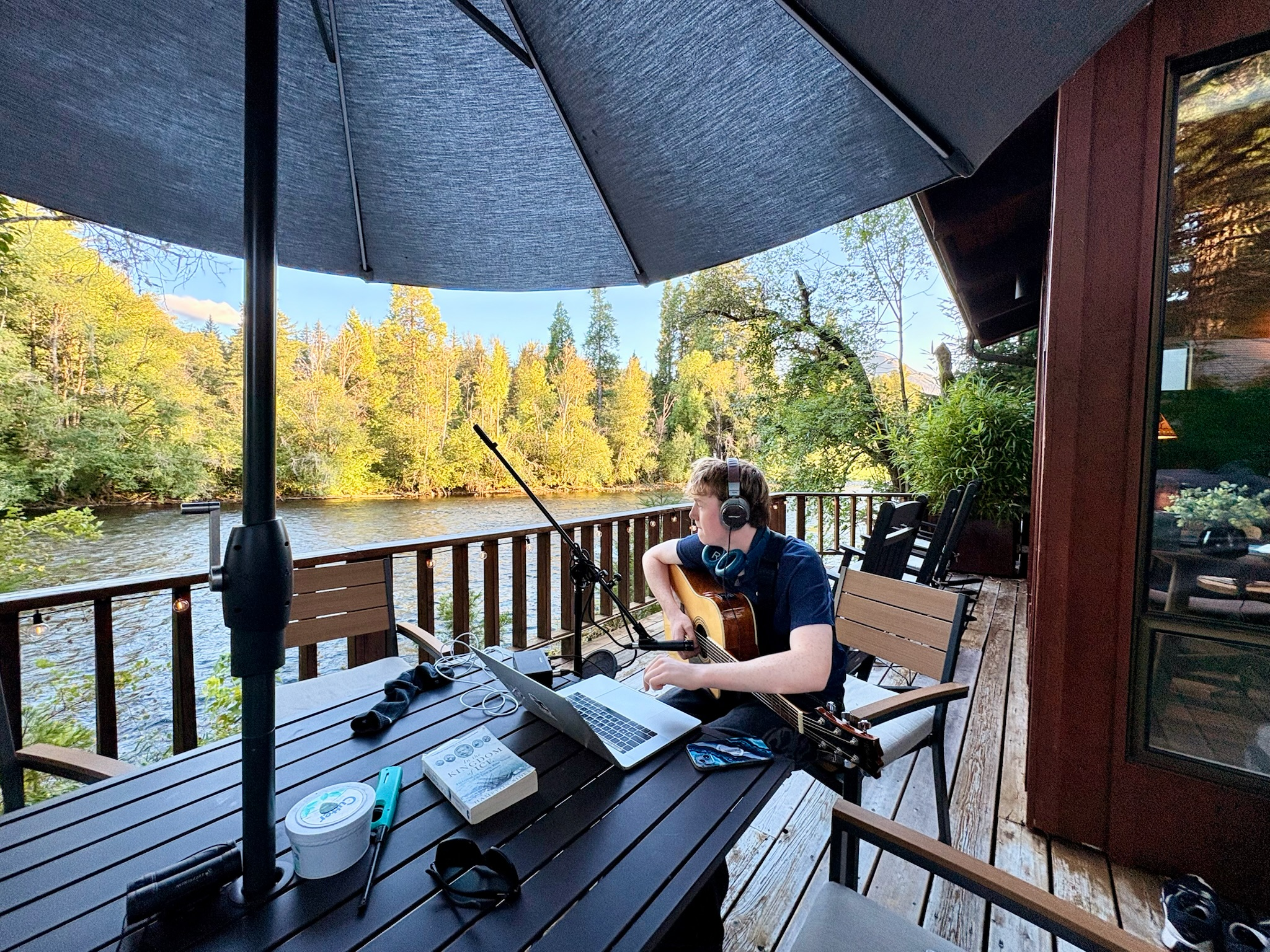 A person wearing headphones sitting on a porch with a guitar, working on a laptop, surrounded by trees and water, shaded by a large umbrella.