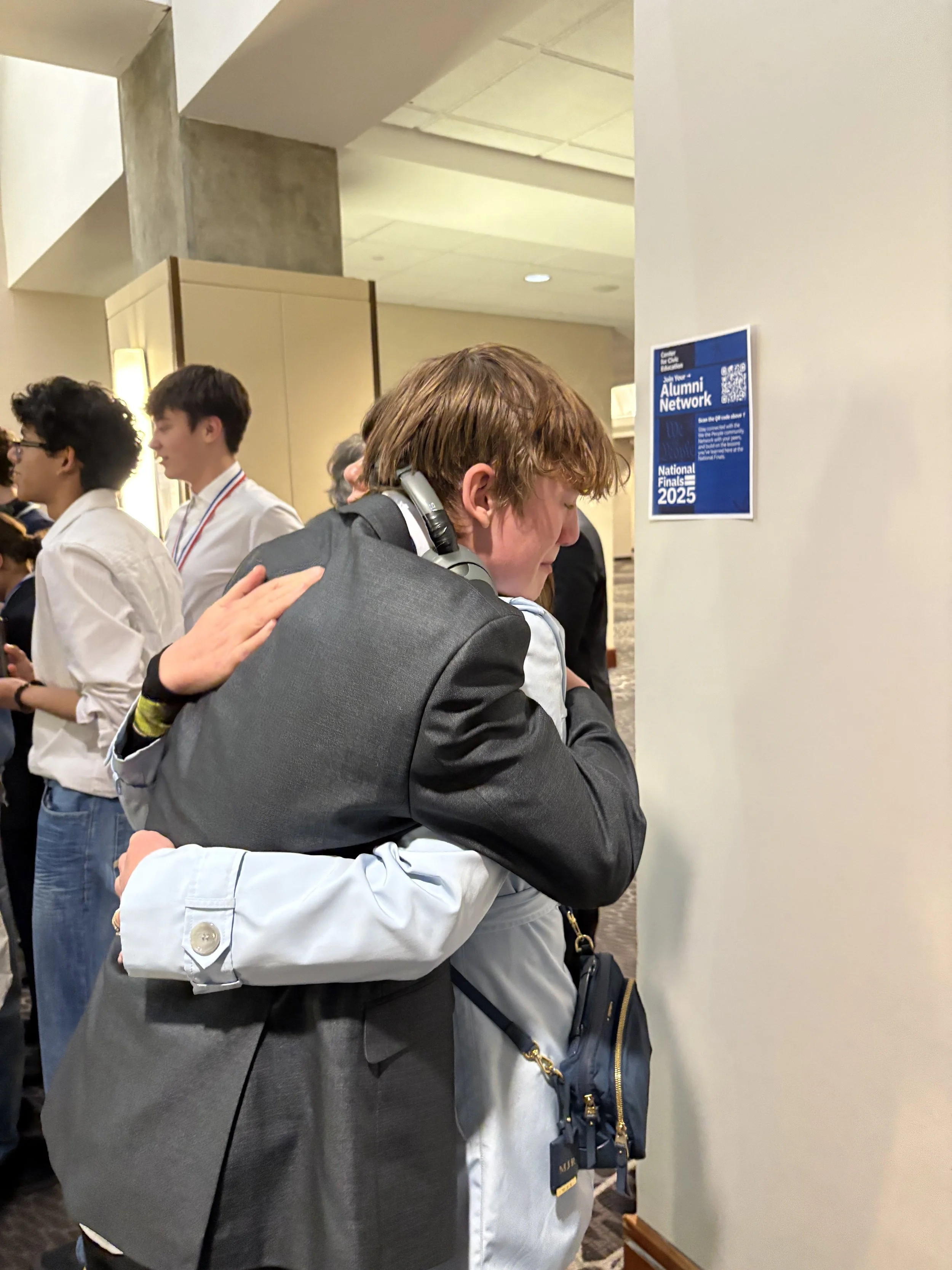 Young man with red hair and headphones, hugging a woman at an event with a sign for the Alumni Network and National Finals 2025 on the wall.