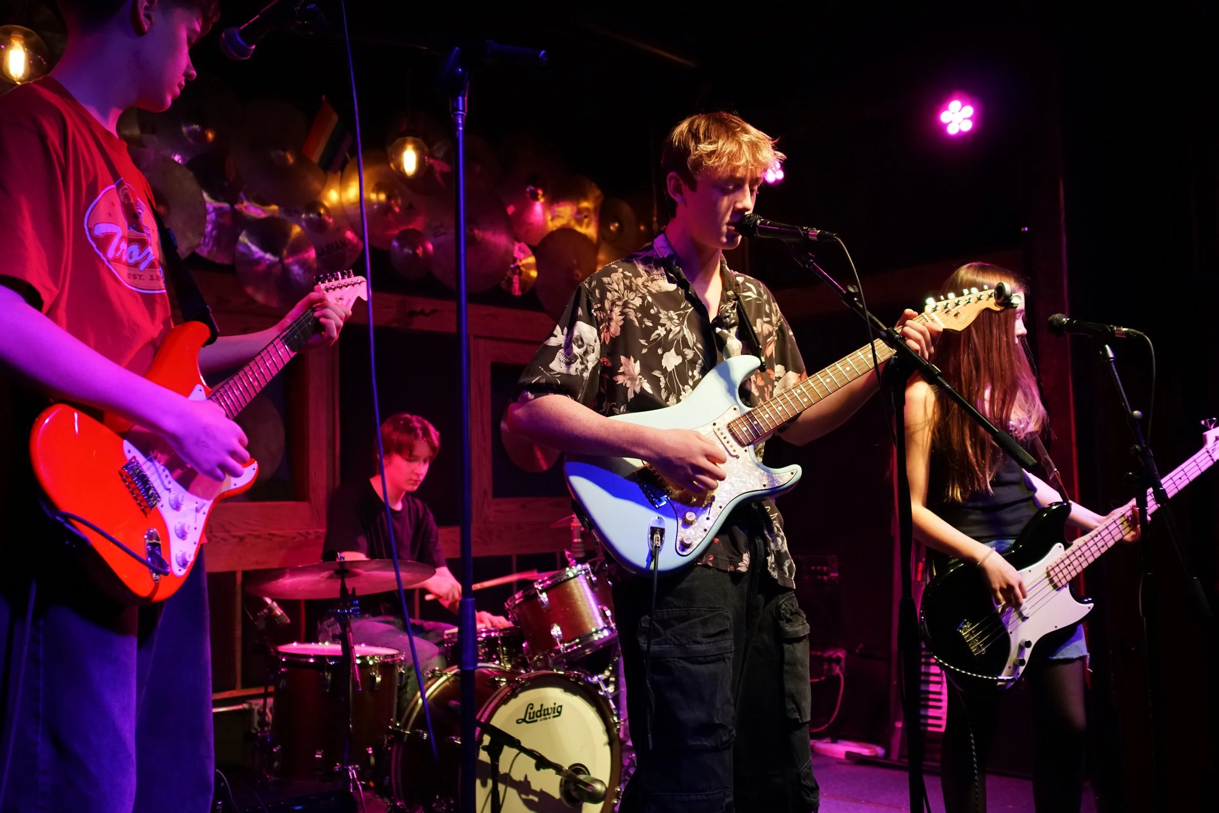 Young band performing on stage with guitars and drums, colorful stage lighting, and a decorated backdrop of metallic spheres.  Lead singer also plays rhythm guitar.