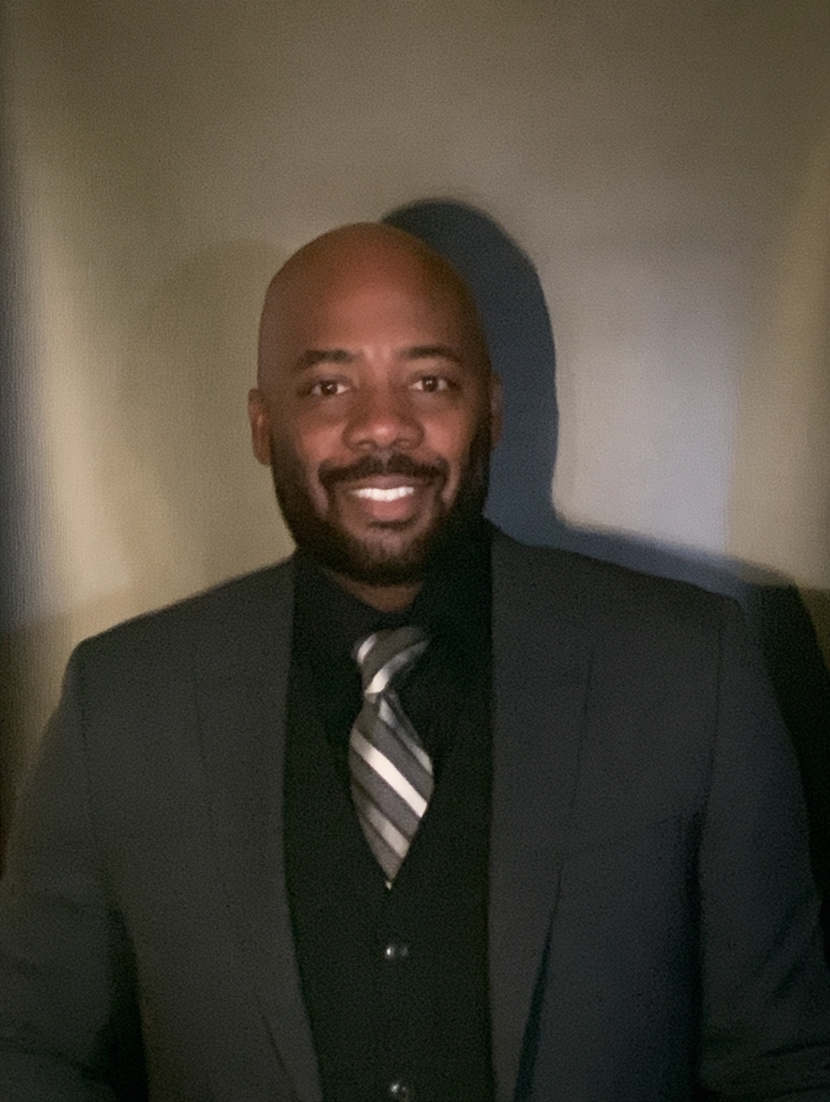 A man with a shaved head and a beard smiling in a black suit, black shirt, and striped gray tie, standing against a neutral wall with a shadow cast behind him.