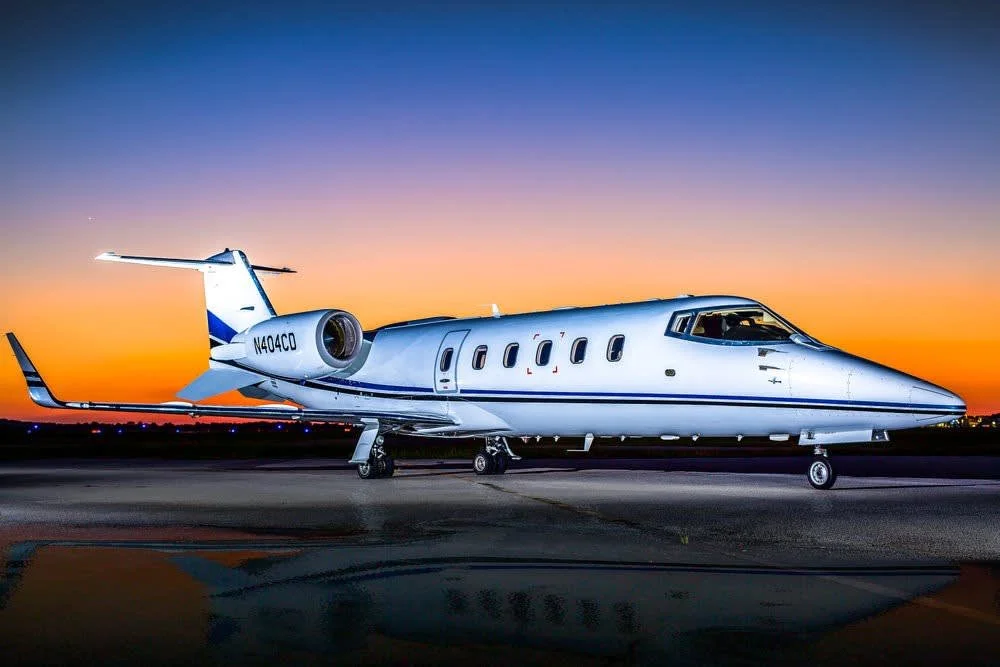 A private jet parked on an airport tarmac during sunset or sunrise with a colorful sky in the background.
