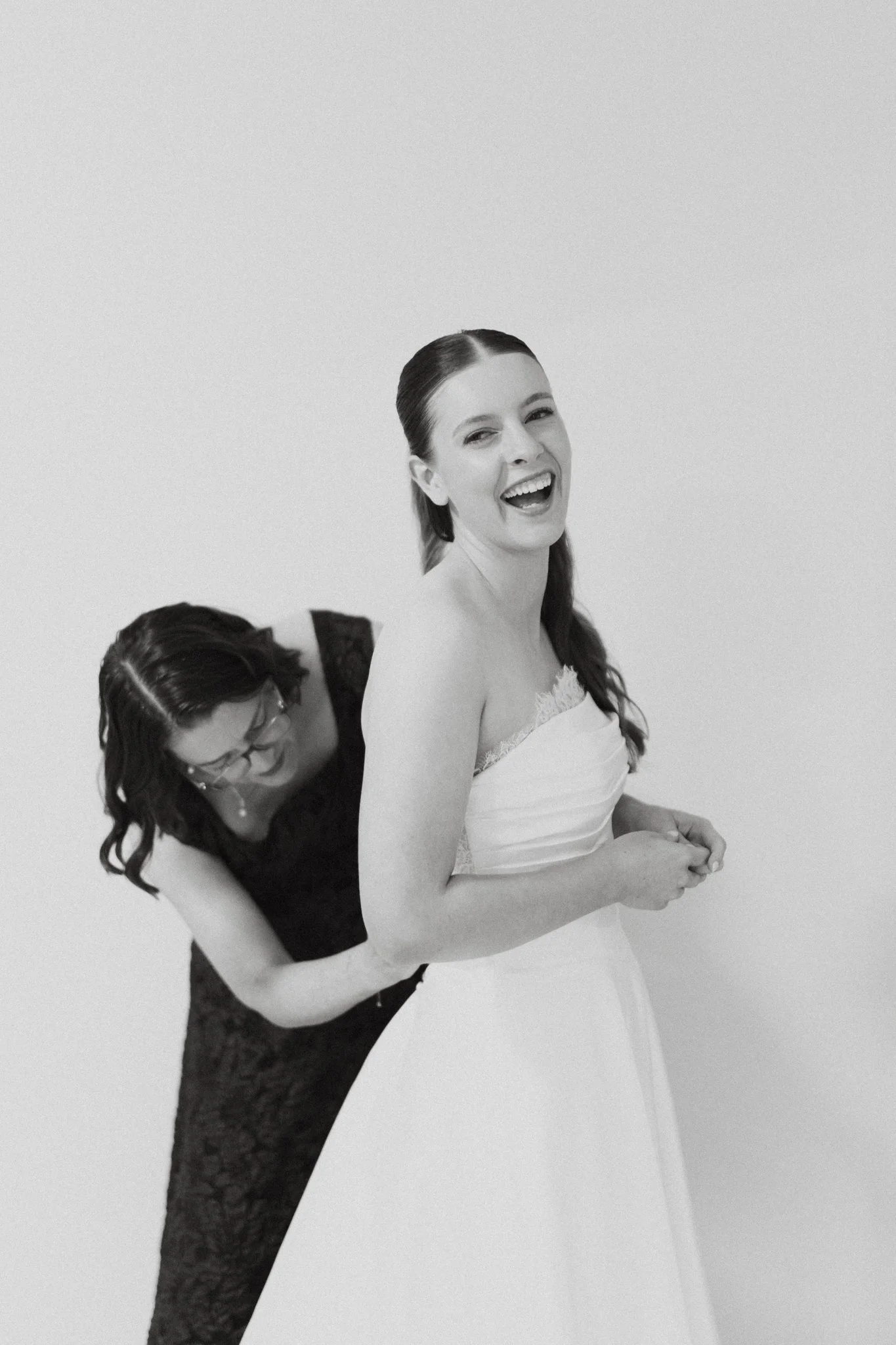 Black and white photo of a laughing bride being helped into her dress