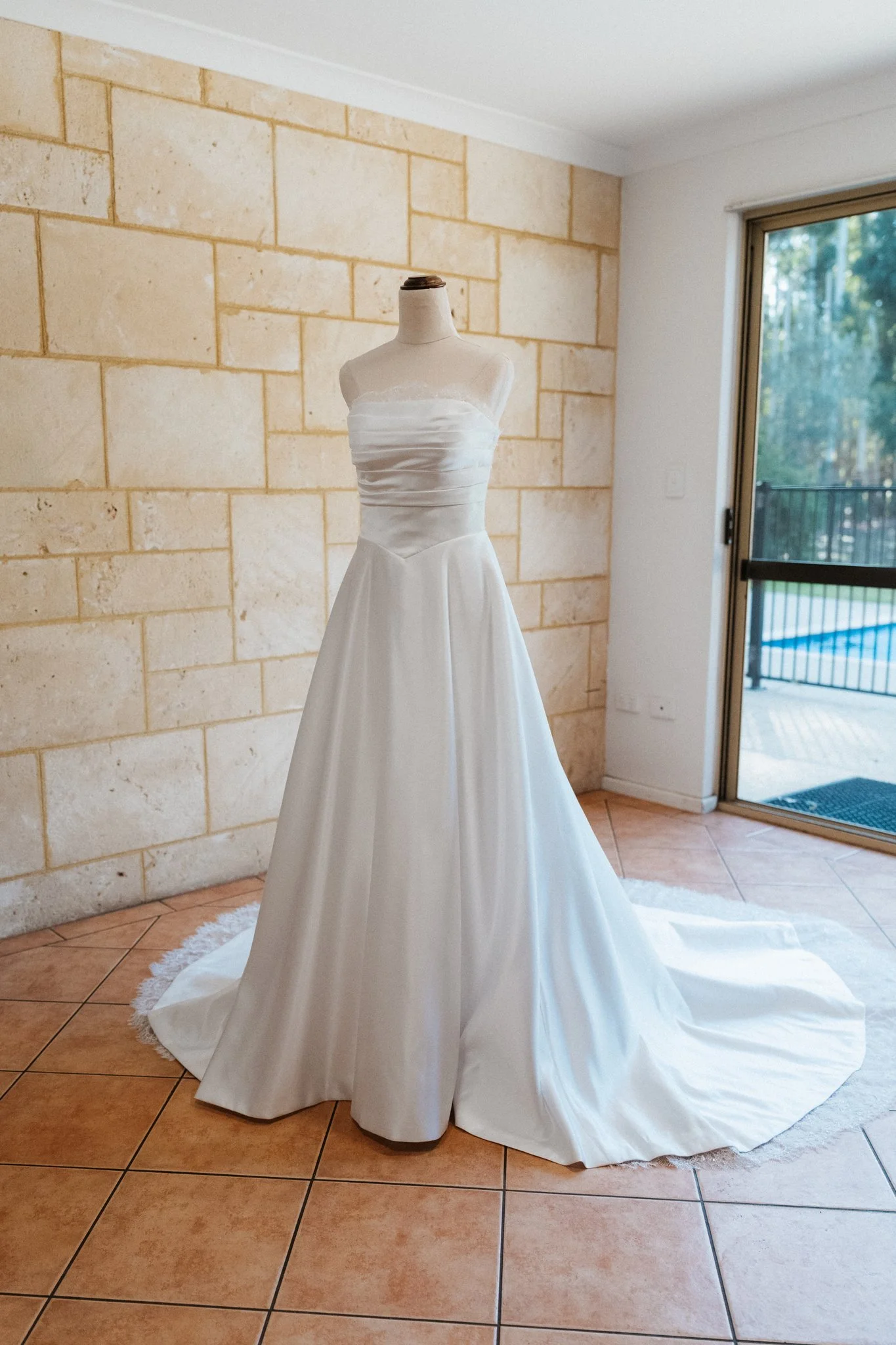 Strapless white wedding gown on a mannequin beside a sandstone wall