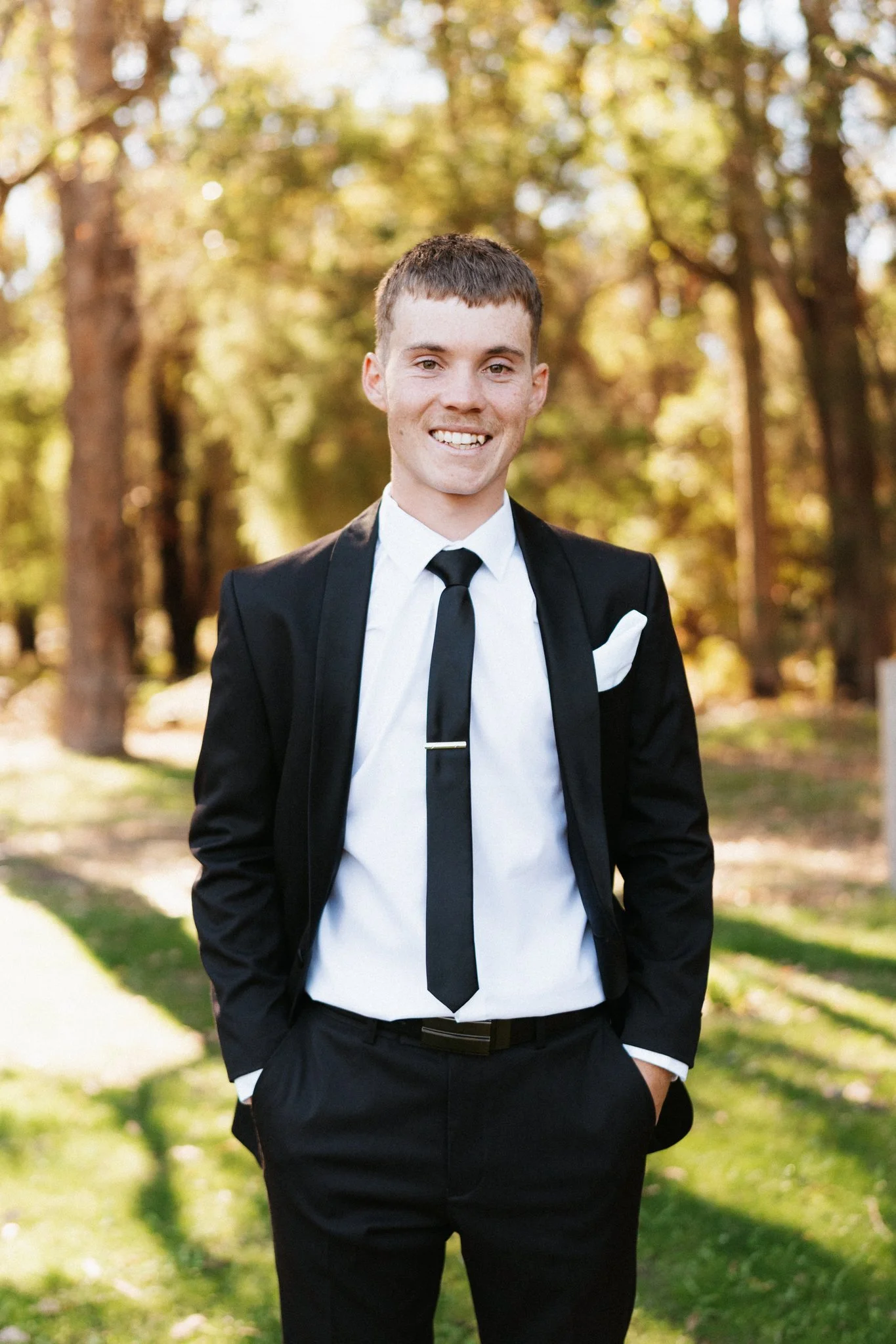 Smiling groom in a black suit and tie standing among bush trees