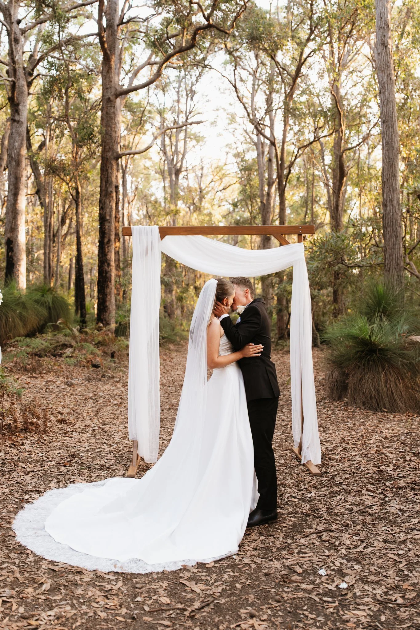Bride and groom standing under a white draped arbour in the forest
