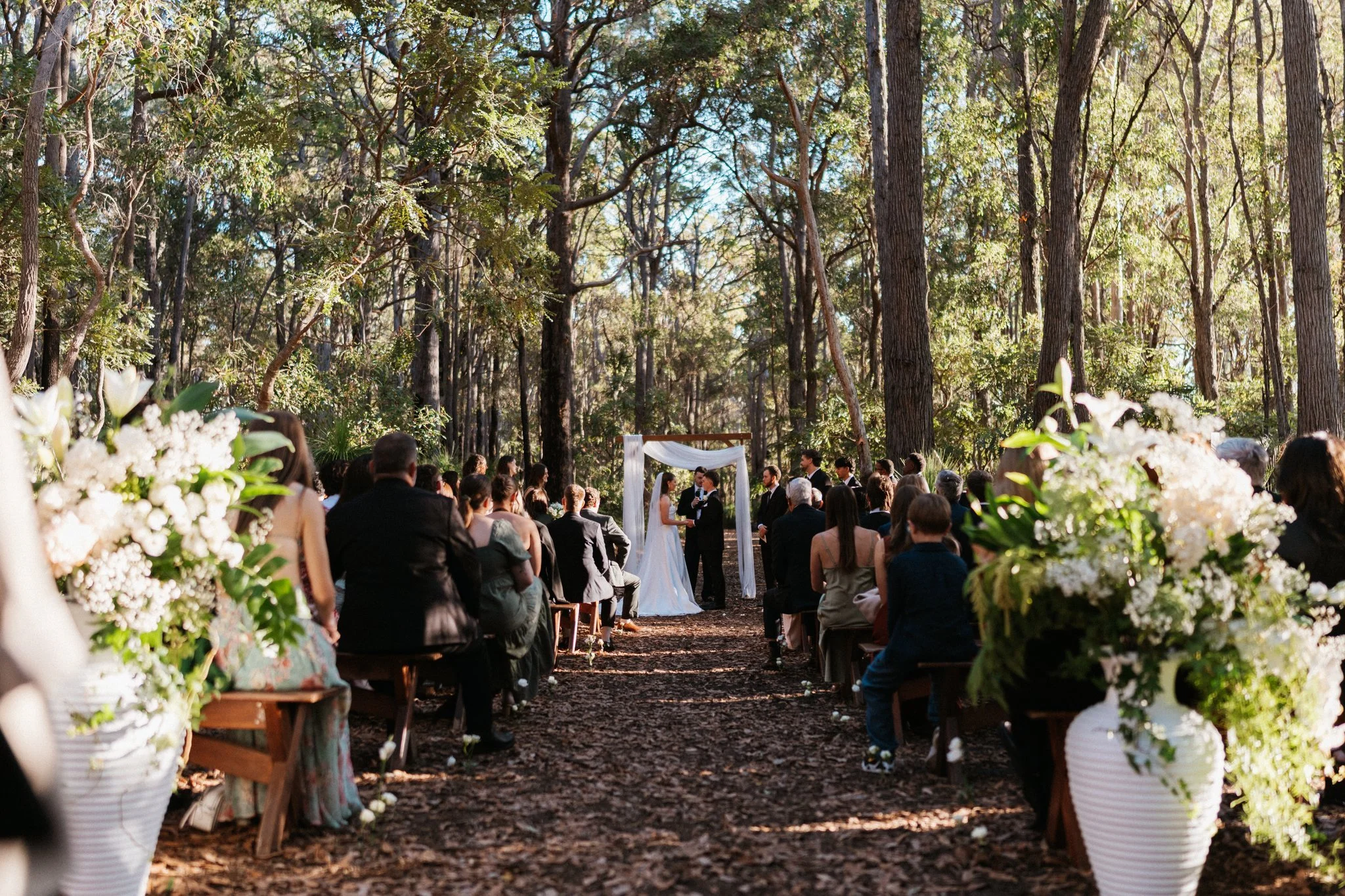 Outdoor ceremony in tall bushland with guests seated along a flower-lined aisle