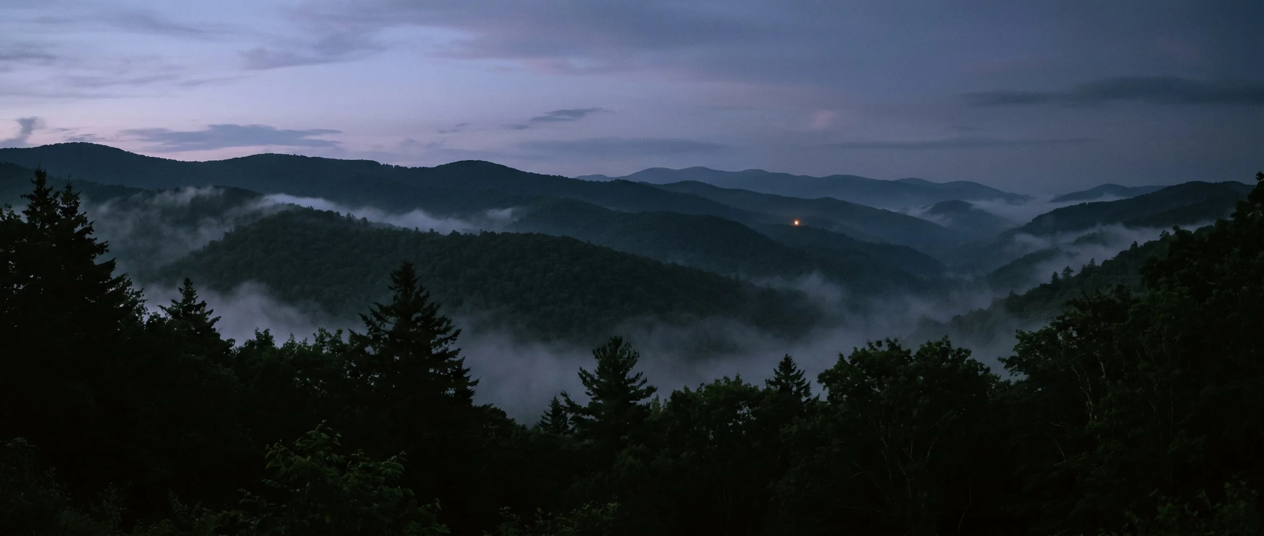 Mountain landscape at dusk with layers of fog rolling through forested ridges and a cloudy sky.