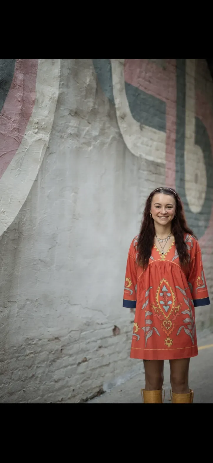 Young woman with long dark hair smiling, wearing a colorful dress and boots, standing against a textured wall with abstract mural art.