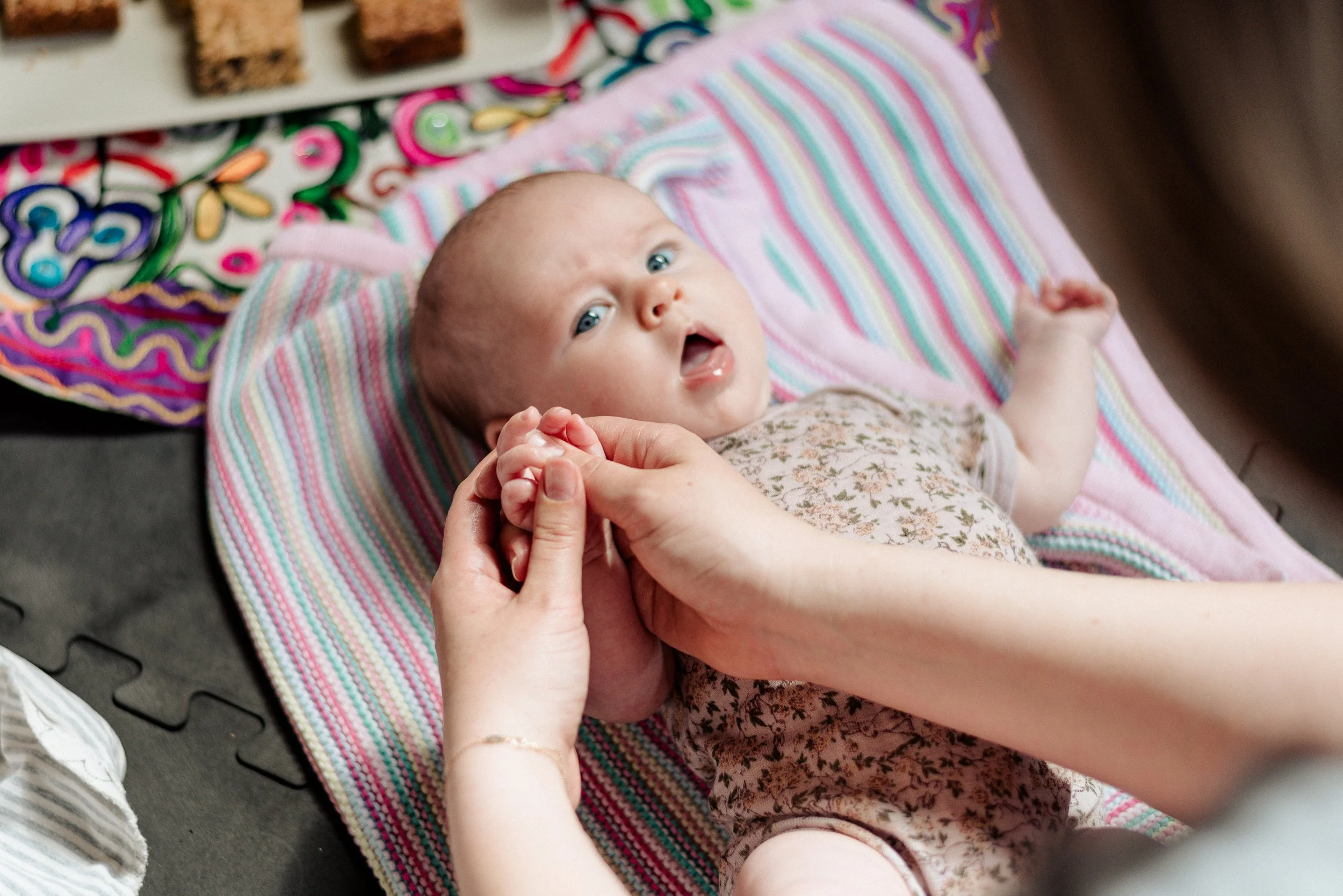 A baby lying on a colorful striped blanket, looking up towards the adult who is giving her a hand massage.