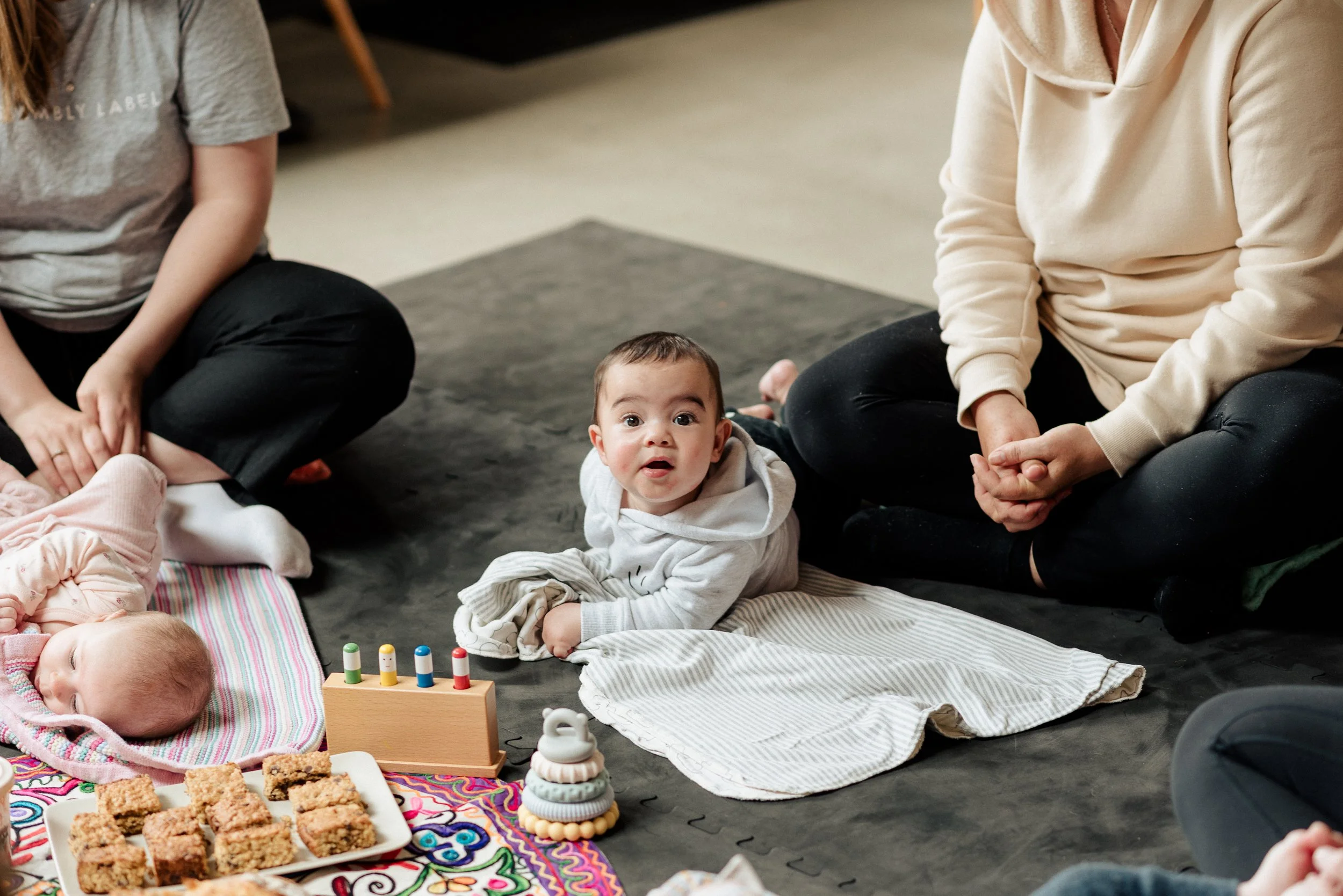 A baby with wide eyes sitting on a blanket surrounded by adults, with snacks and toys nearby.