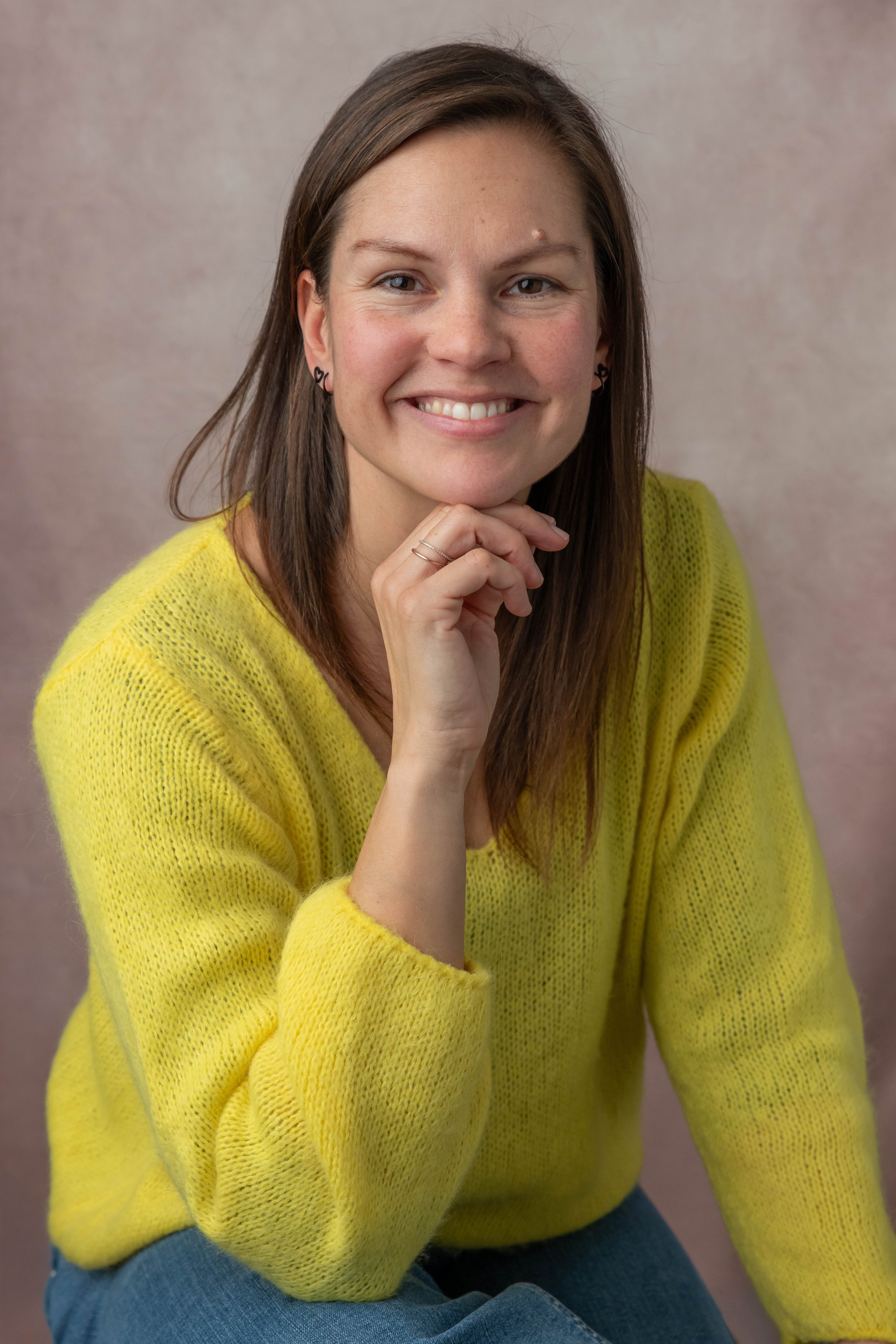 A young woman with long brown hair and light skin. She is smiling, resting her chin on her right hand. She is wearing a yellow sweater and blue jeans, against a plain muted background.