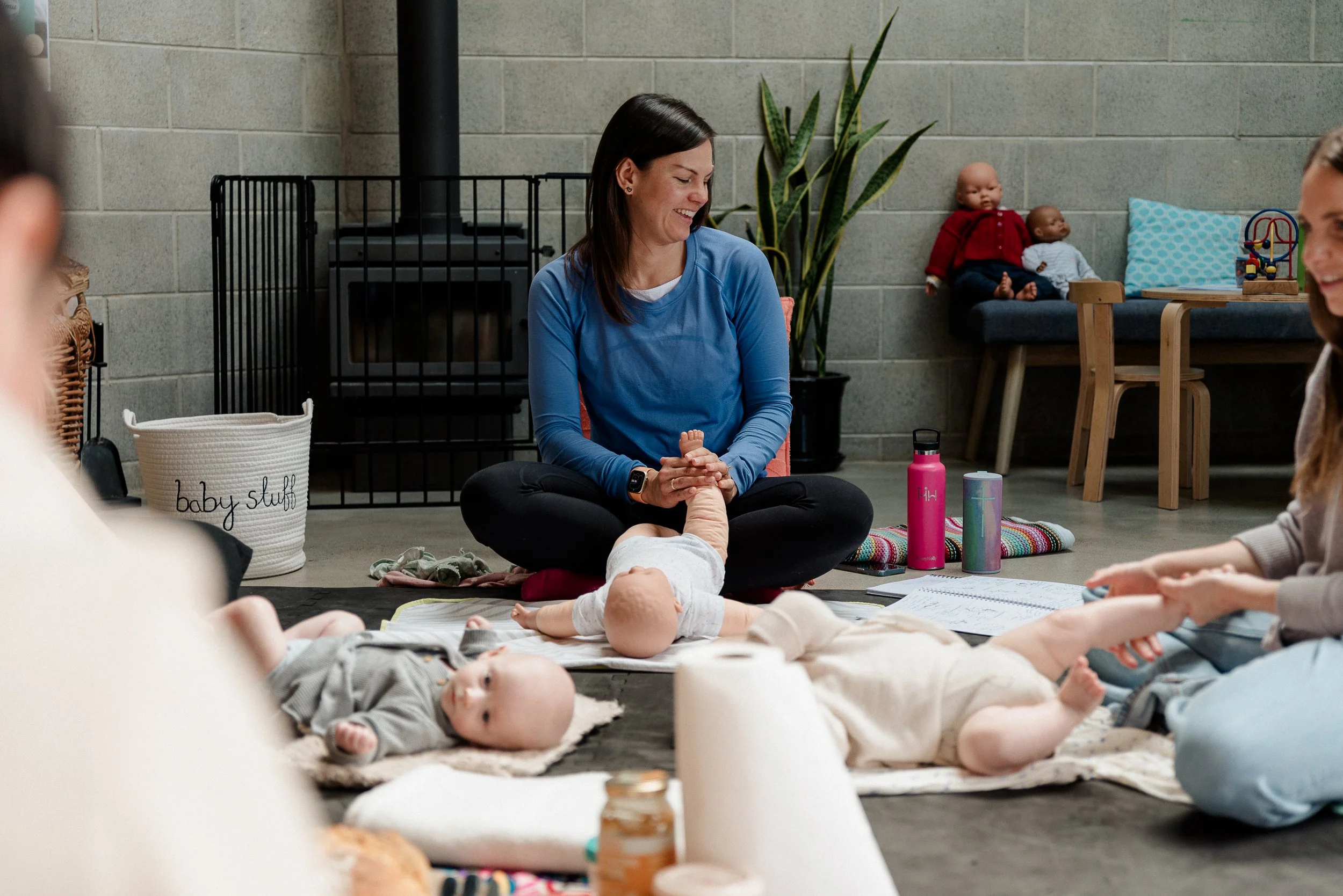 A white woman with brown hair smiling and sitting cross-legged on the floor during a baby massage class, surrounded by babies and caregivers, with toys nearby, in a room with a grey brick wall, a large plant, and seating.