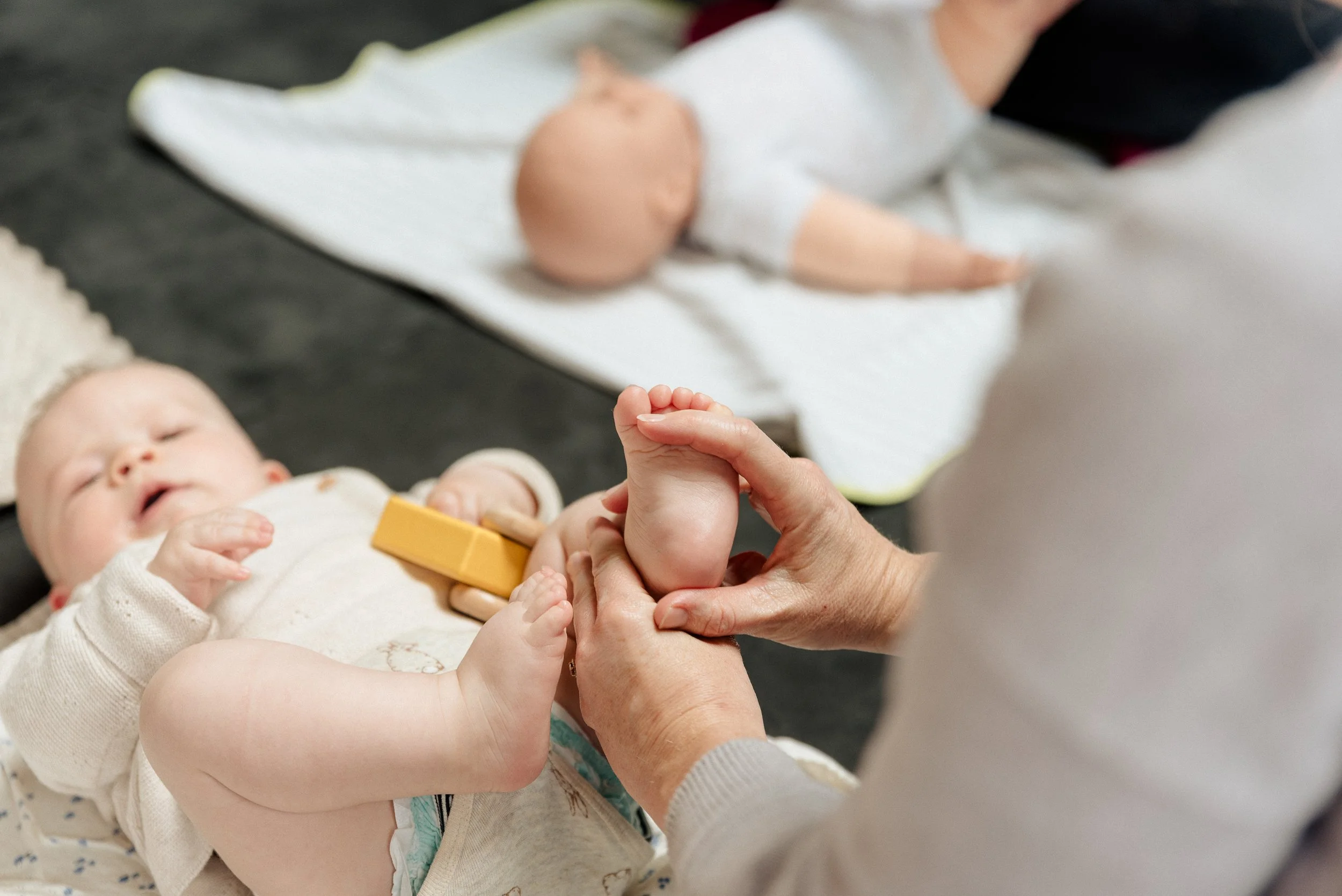 A baby lying on a soft surface receiving a foot massage with its caregiver's hands visible. A baby massage demonstration doll can be seen in the background.
