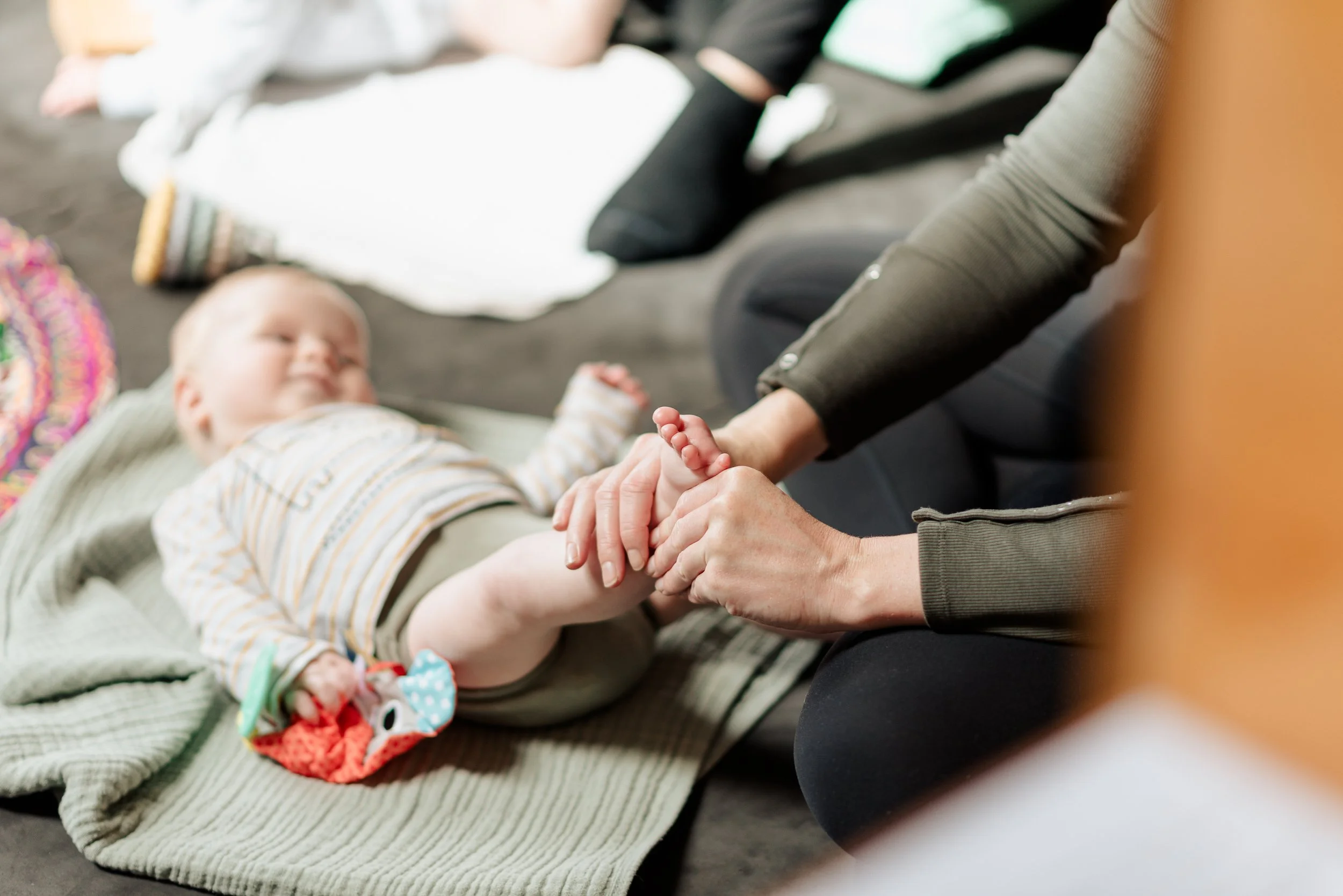 A young child lying on a blanket with a colourful toy, receiving a leg massage from an adult in a grey sweater.