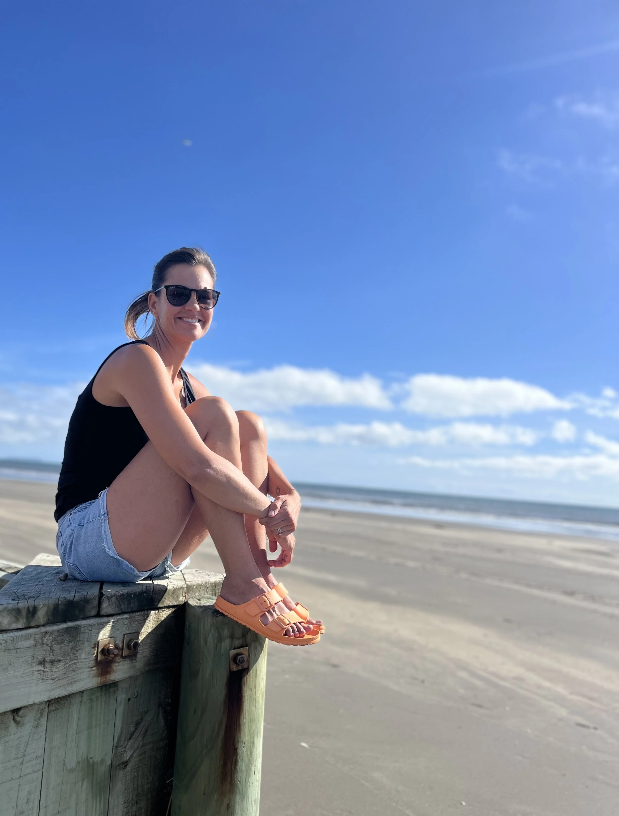 A caucasian woman sitting on a wooden pier at the beach, smiling, wearing sunglasses, a black tank top, denim shorts, and orange sandals, with a clear blue sky and ocean in the background.