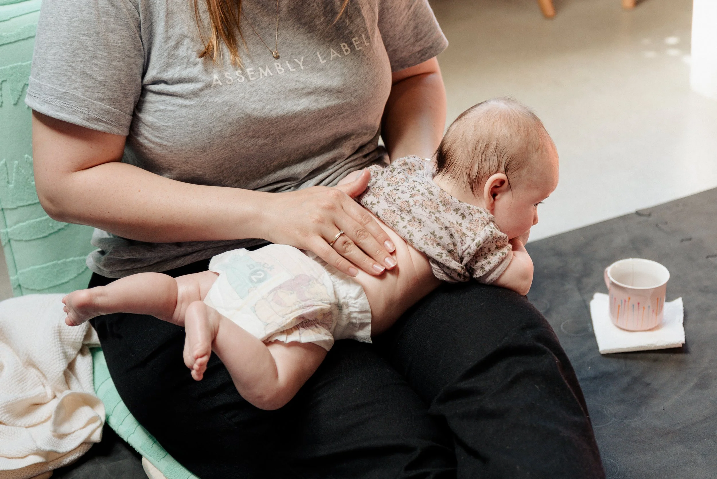 A woman with reddish hair wearing a gray T-shirt and black pants is seated, holding a young baby on her lap. The baby, dressed in a floral outfit and a diaper, is lying face down on her lap with feet hanging over her side. The woman is gently touchin